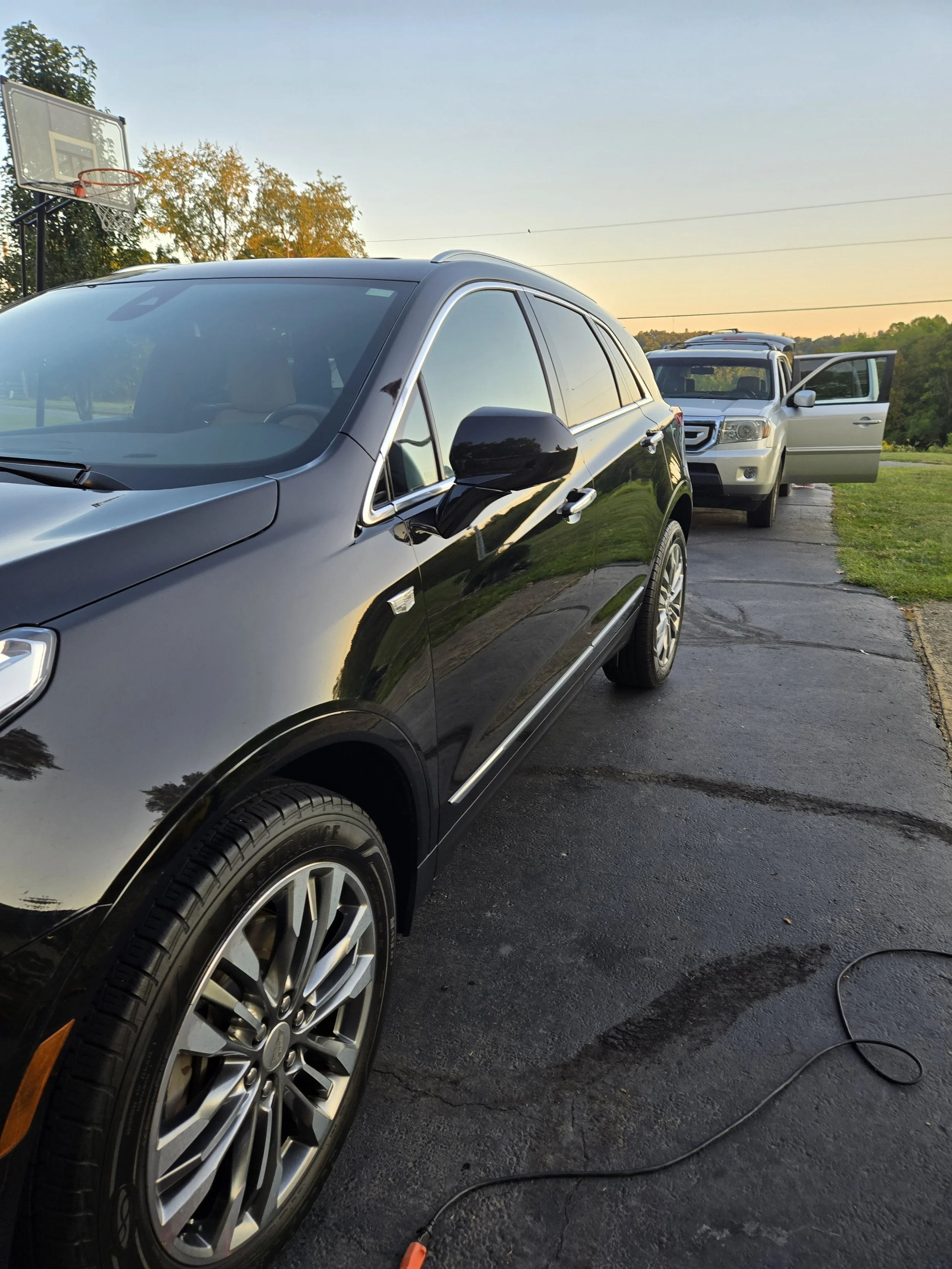 Black SUV parked on driveway with silver SUV in background, open rear passenger door, outdoor setting during sunset, basketball hoop nearby.