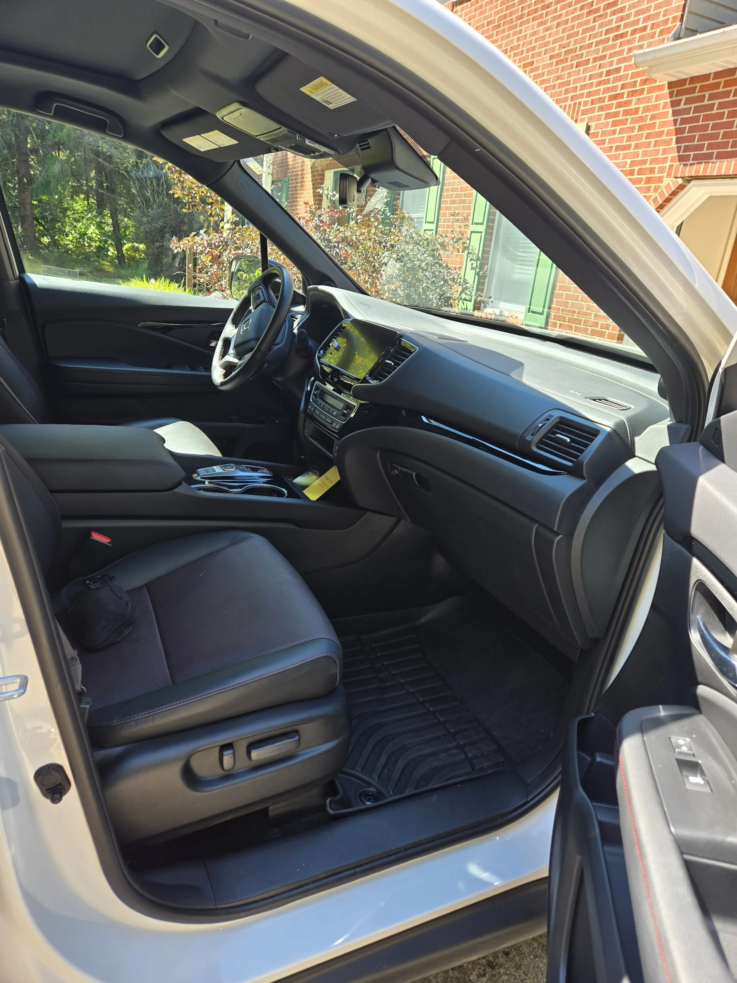 Interior of a modern car showing the front passenger seat, dashboard, steering wheel, and center console, with a view through the windshield of a brick house and greenery outside.