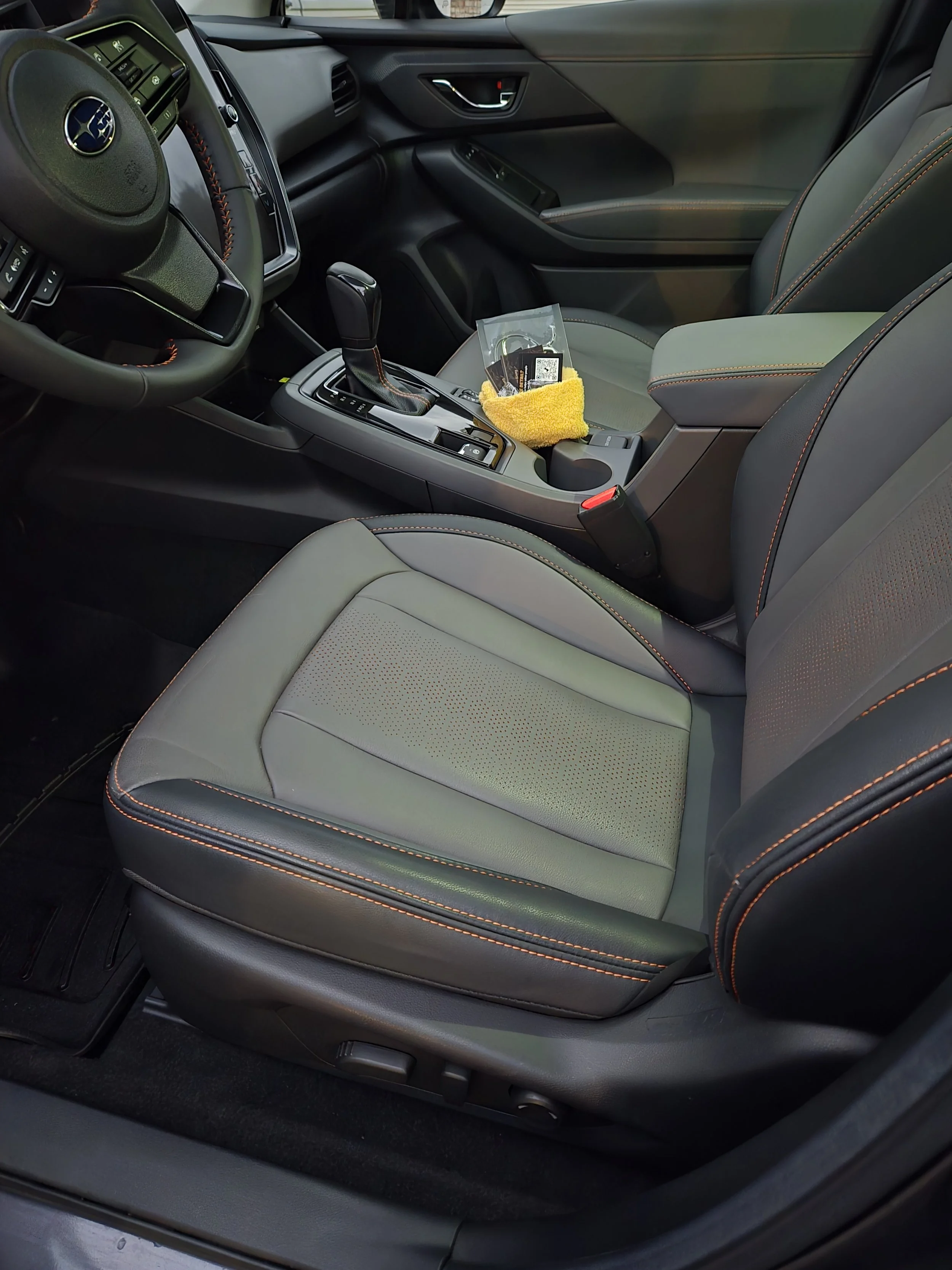 Interior of a car showing the front passenger seat, center console with gear shift, dashboard, and steering wheel with Subaru logo. Items on the center console include a yellow cloth, some small objects, and a plastic bag.