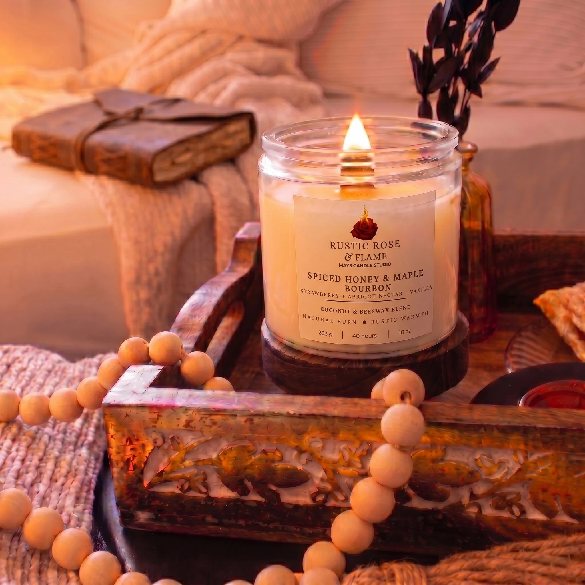 A lit candle labeled 'Rustic Rose & Flame' with scent notes of spiced honey, maple bourbon, strawberry, apricot nectar, vanilla, coconut, beeswax, and warm notes. The candle is on a wooden surface, surrounded by a wooden bead garland, with a blurred rustic background.
