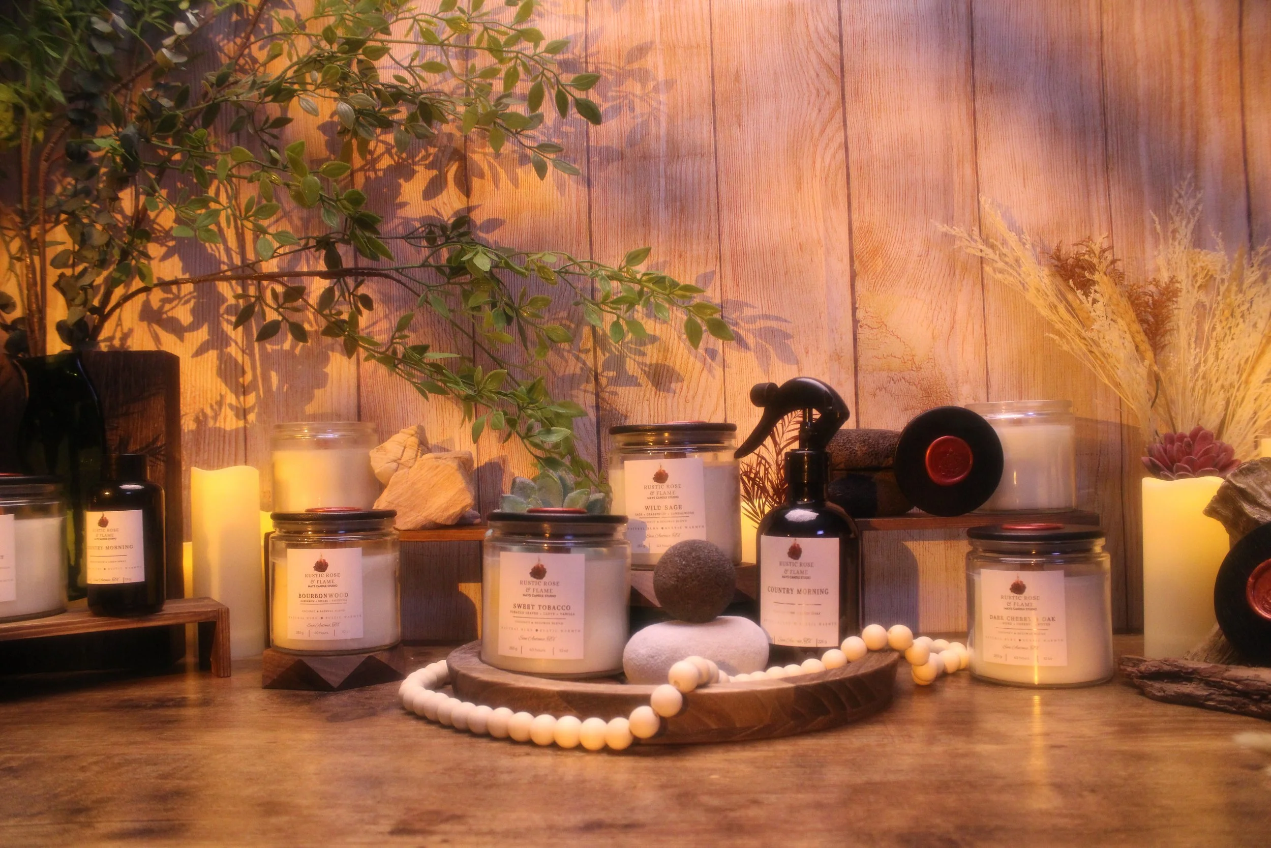 A wooden shelf displaying candles, essential oils, and natural decorative items, with a leafy plant and dried grasses in the background, illuminated with soft warm lighting.