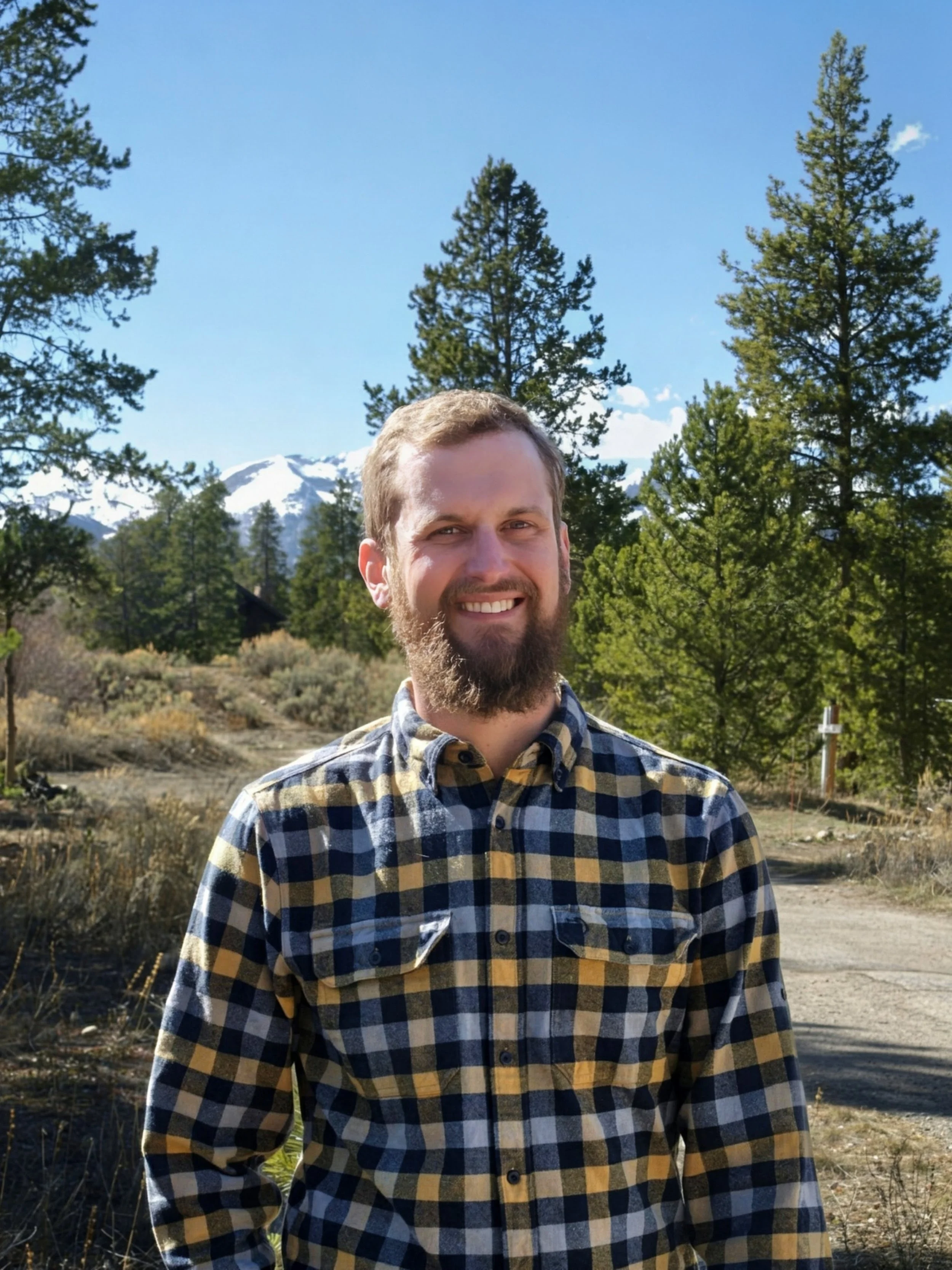 Smiling man with a beard in a plaid shirt standing outdoors with trees, mountains, and a clear blue sky in the background.