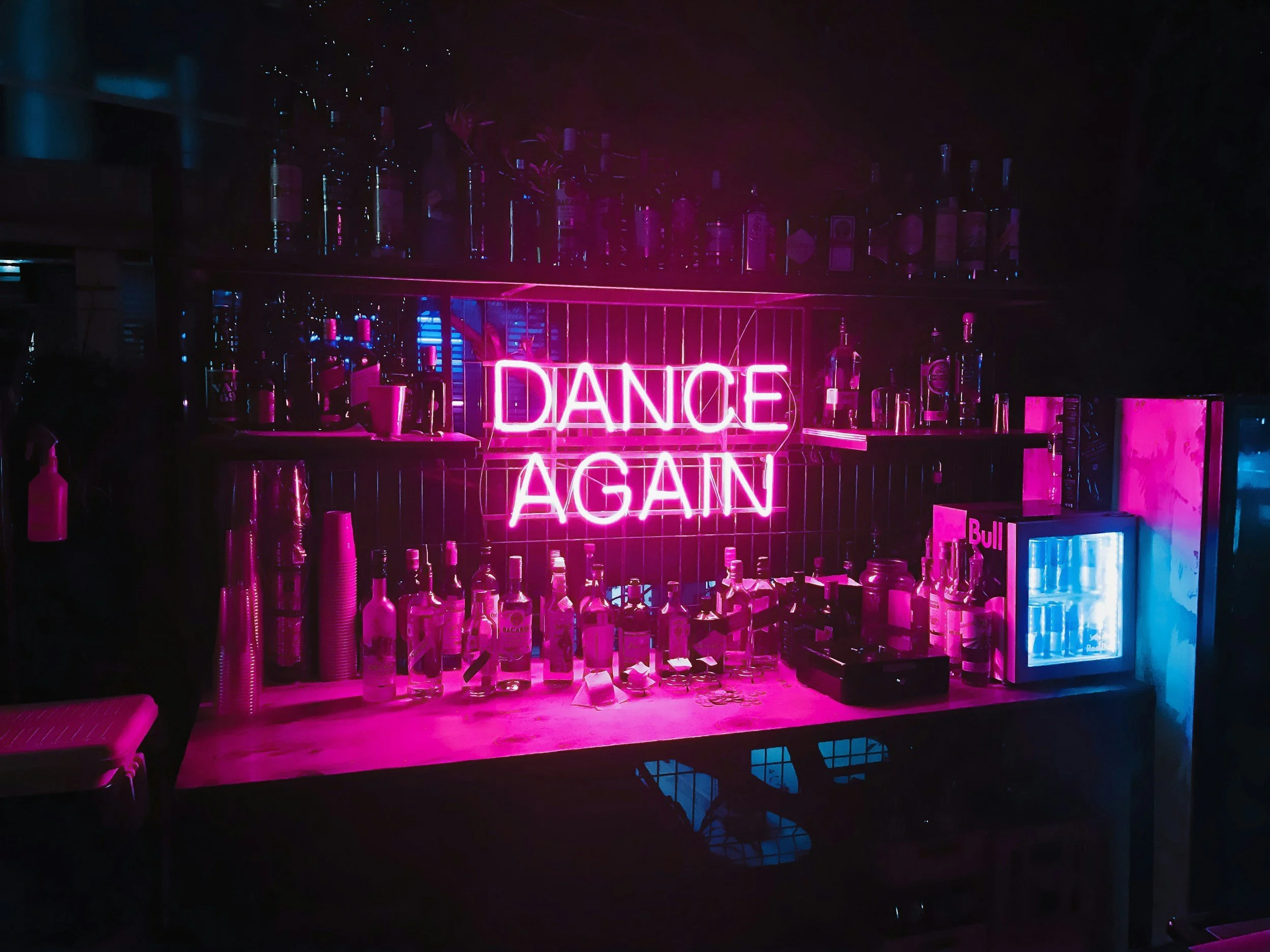 A neon pink sign that says 'DANCE AGAIN' hangs above a bar with bottles of alcohol and bar equipment, illuminated by pink and blue lighting.