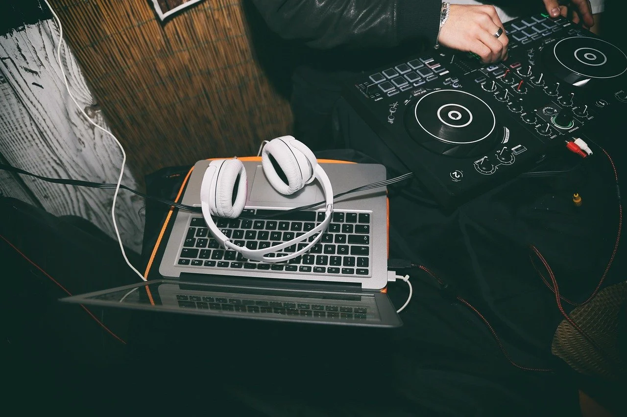 DJ mixing equipment, a laptop with white headphones resting on it, and a person's hand operating the DJ controller.