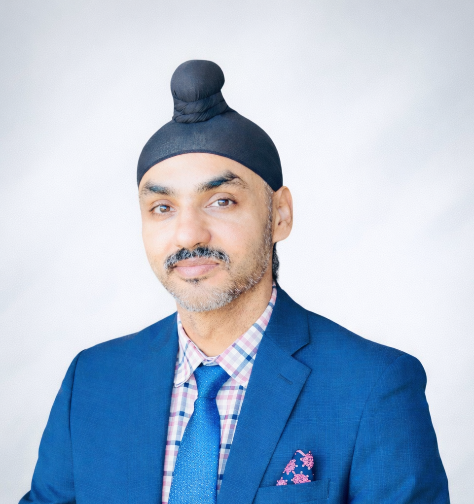 A man wearing a blue suit, checkered shirt, and matching tie with a pink pocket square, also wearing a black head covering, against a plain white background.