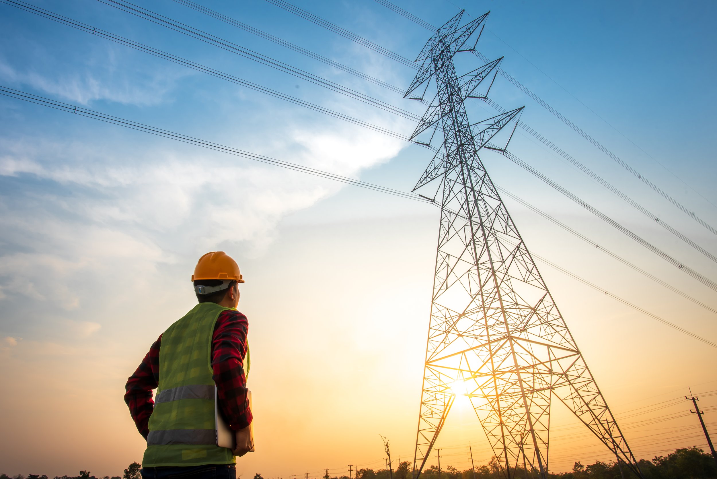 A worker wearing a yellow safety helmet and green safety vest standing outdoors and looking at a tall electrical transmission tower during sunset or sunrise.