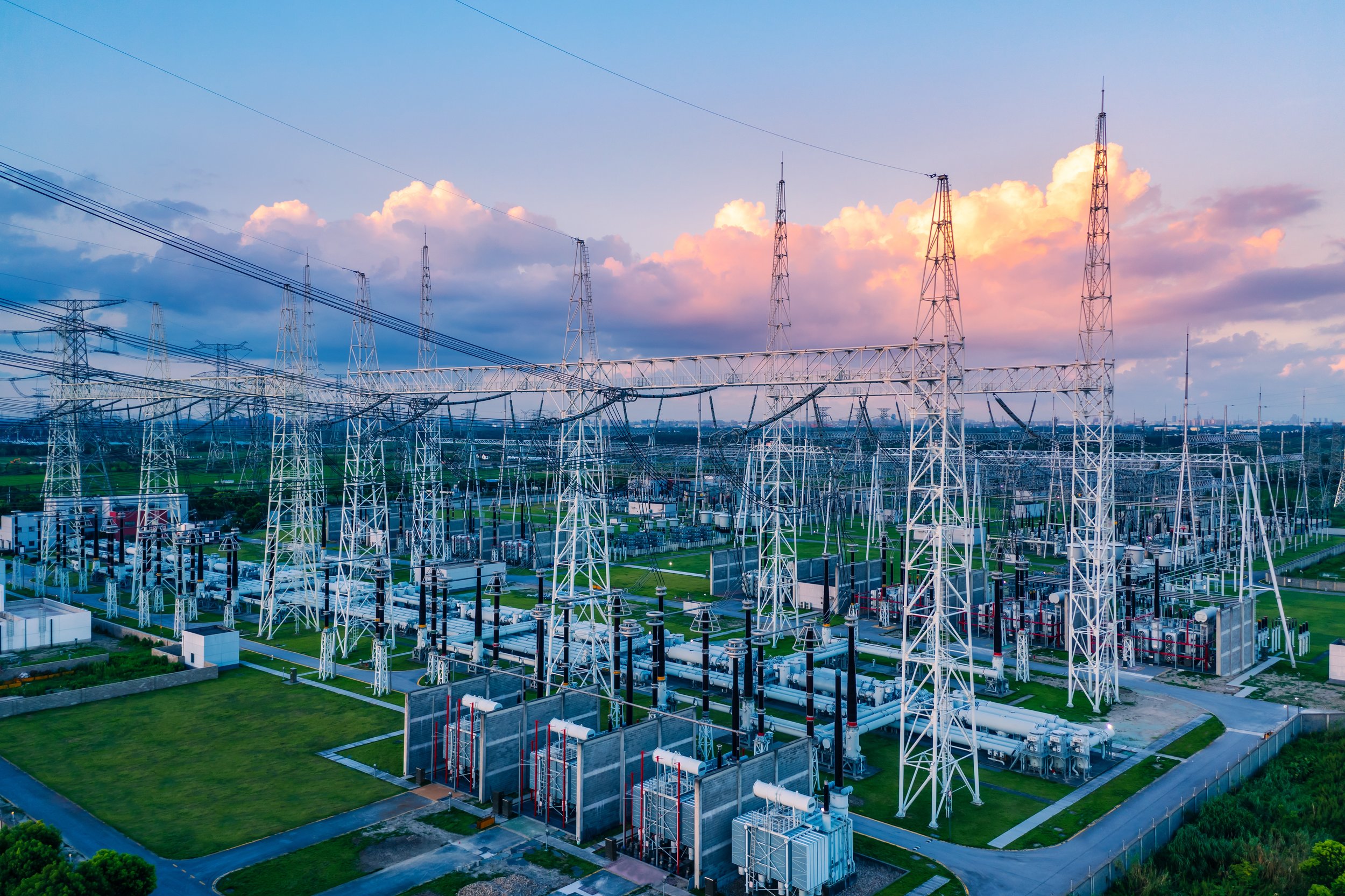 An electrical substation with multiple transmission towers, power lines, and equipment set against a sky with clouds at sunset.
