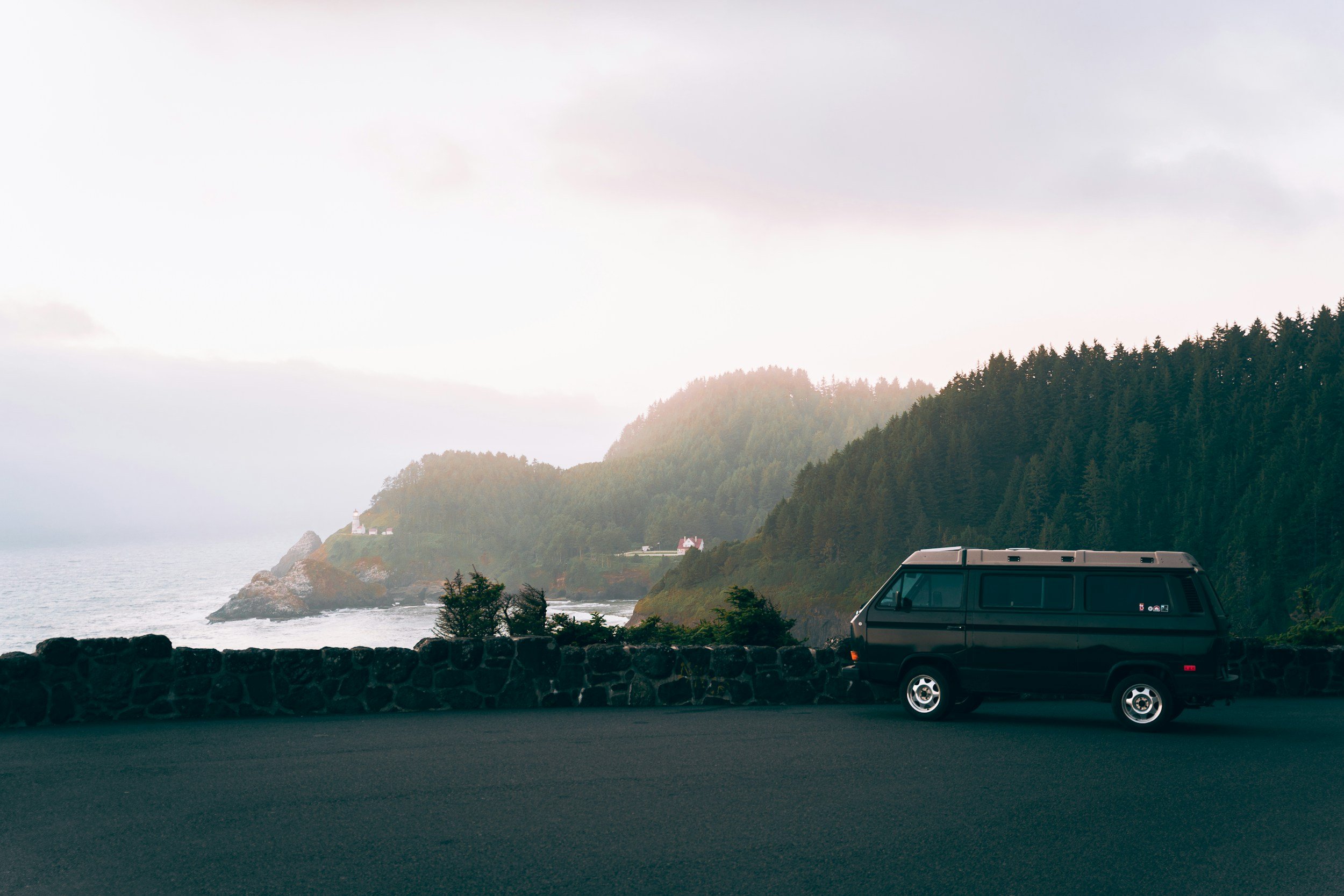 A black van parked near a stone barrier on a road overlooking the ocean with forested hills in the background and cloudy sky.
