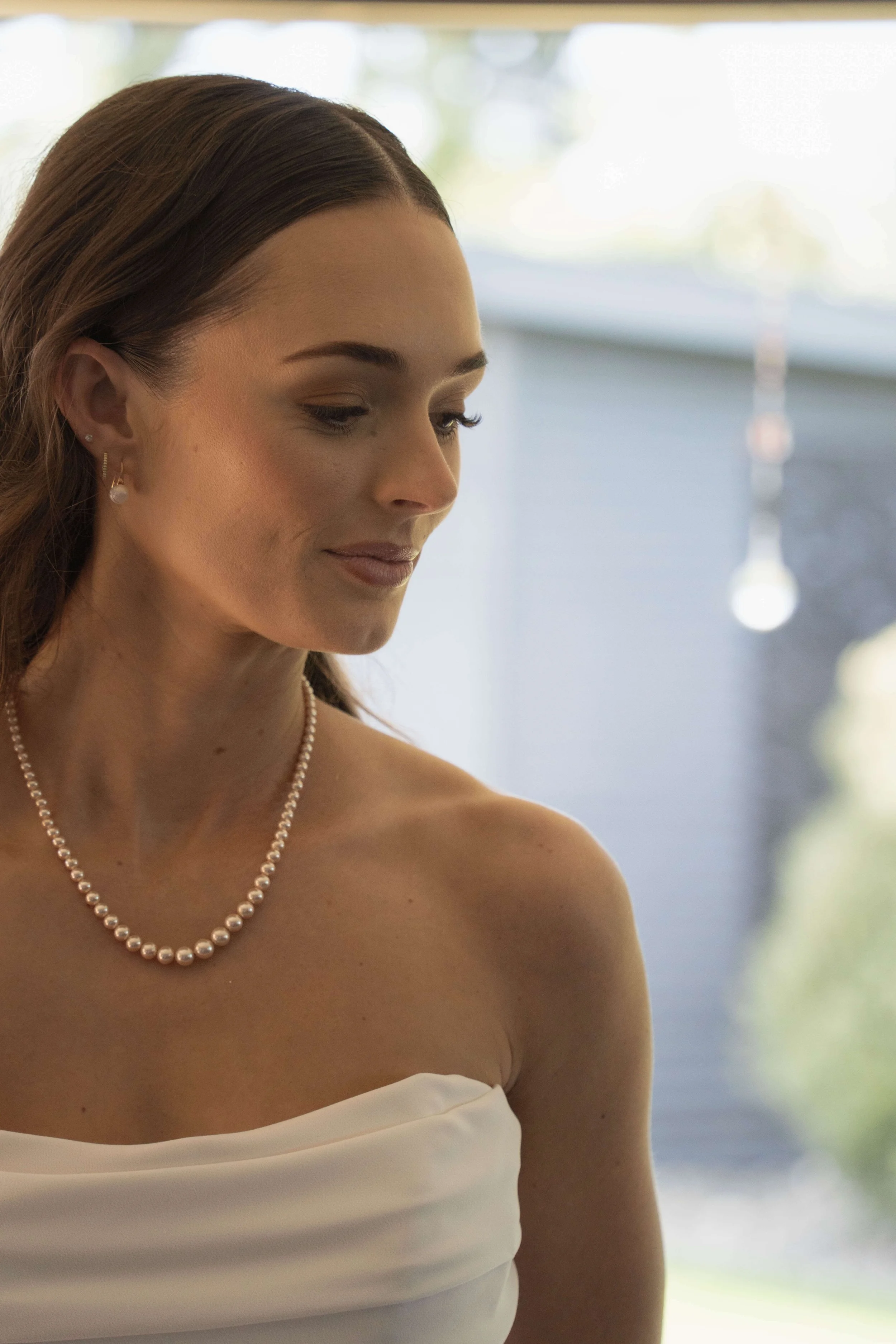 A close-up of a woman in a white strapless dress with pearl jewelry, standing indoors with sunlight coming through a window.