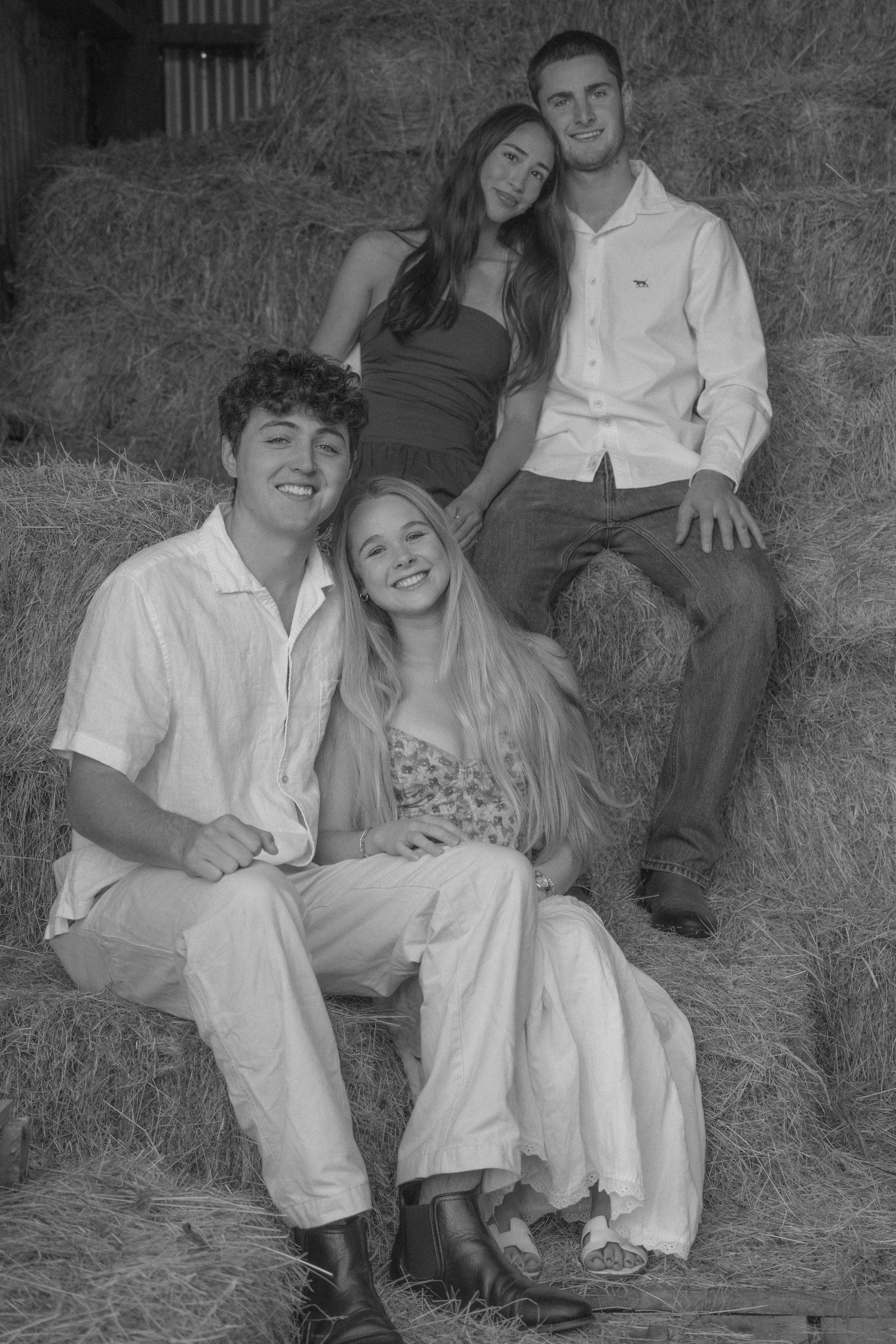 Group of five young people sitting on hay bales in a barn or farm setting, smiling and posing for the camera.