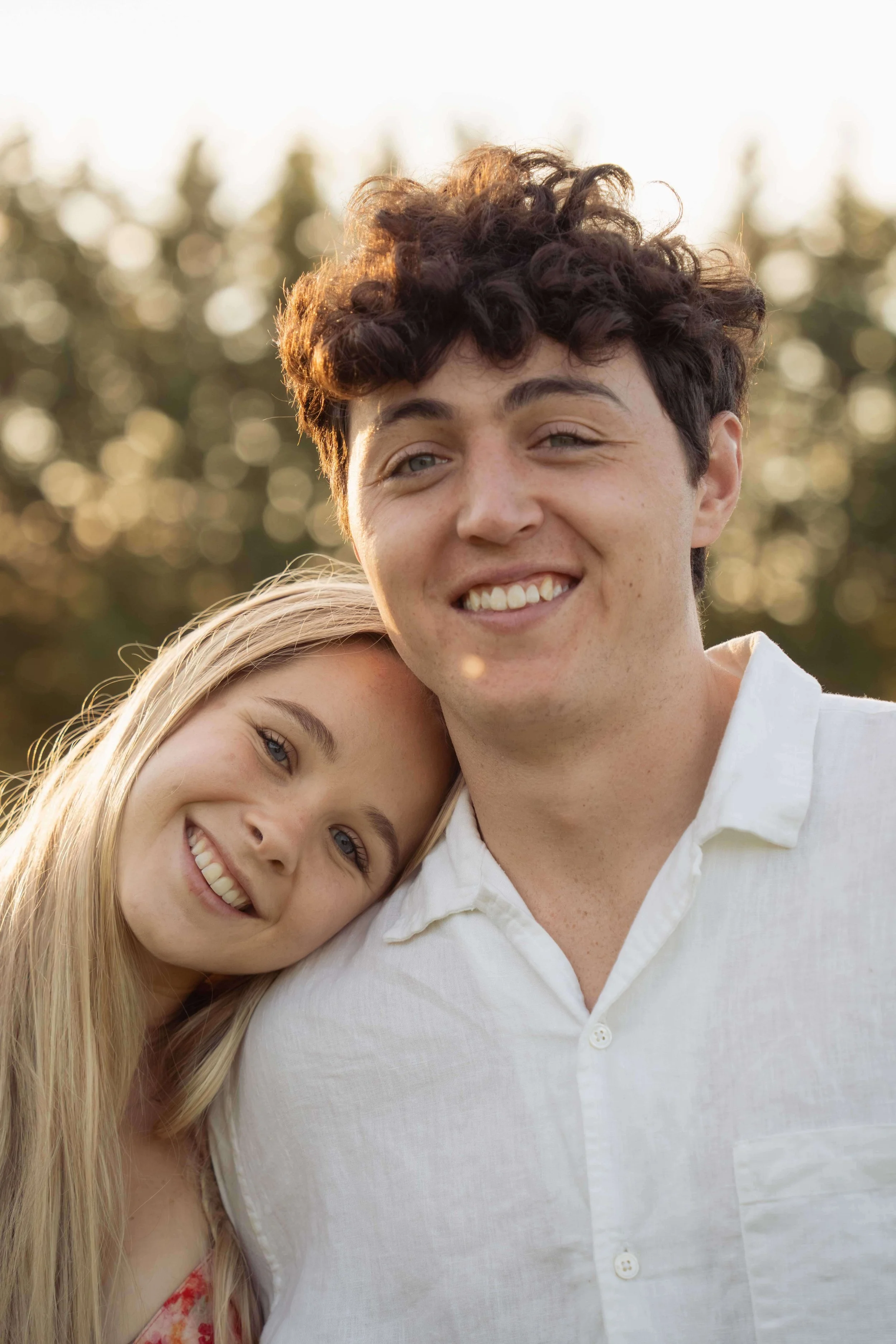 A young woman leaning her head on a young man's shoulder, both smiling, outdoors during golden hour.