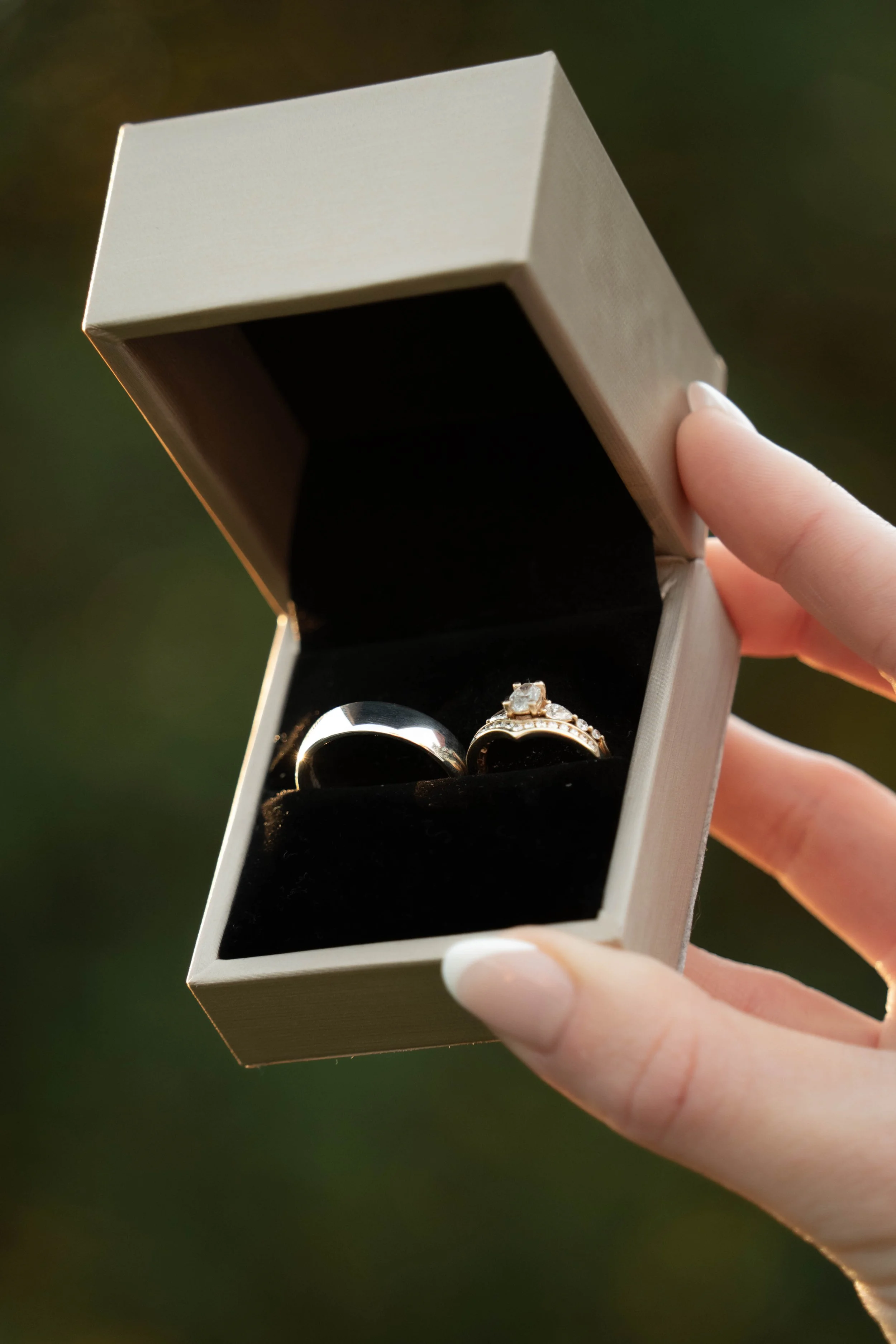 A hand holding an open jewelry box with two rings inside, one is a plain silver band and the other is an engagement ring with a large stone surrounded by smaller stones.