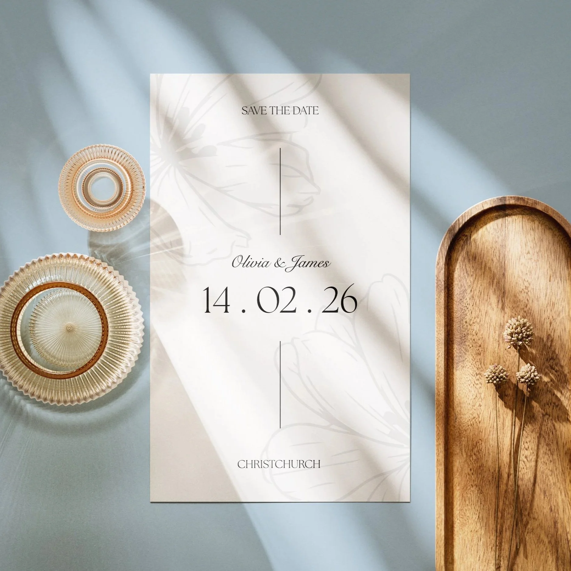 A wedding save-the-date card with the date 14.02.26, names Olivia and James, and location Christchurch, placed on a pastel blue surface with two decorative glass coasters and a wooden tray with small dried flowers.