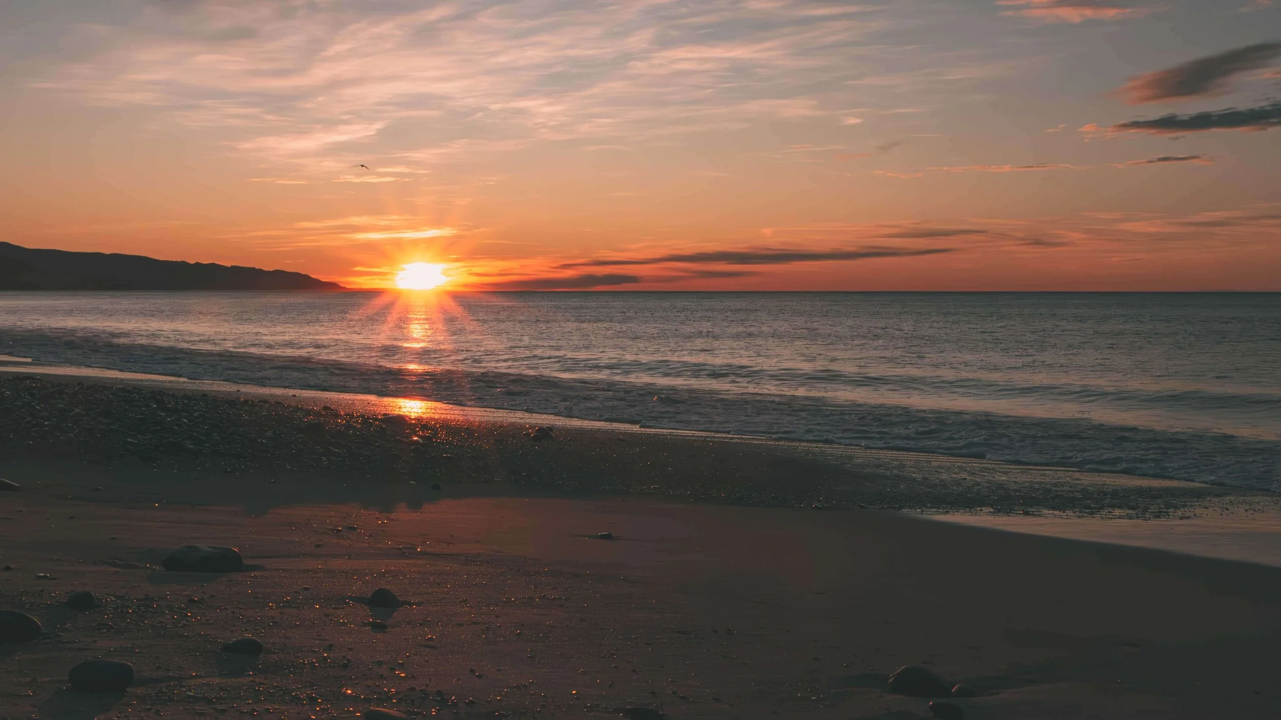 Sunset over the ocean with a partly cloudy sky, a mountain range in the distance, and pebbles on the sandy beach in the foreground.