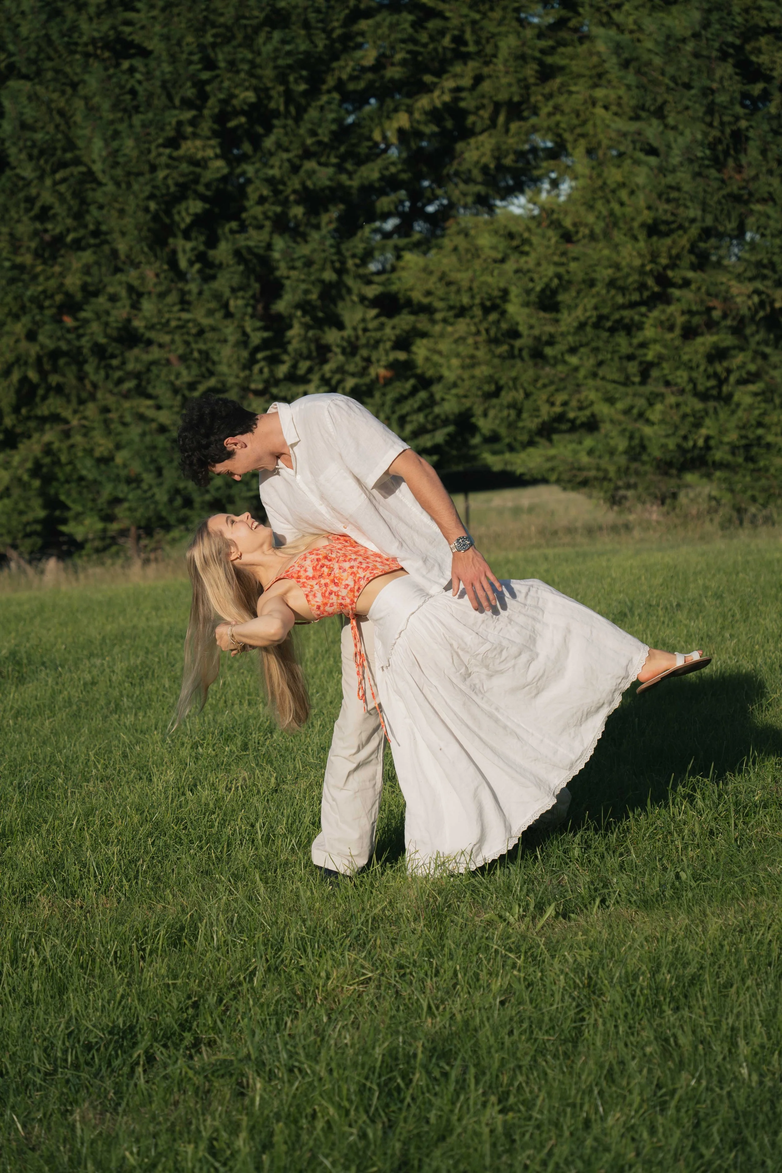 A man and woman dancing outdoors on a grassy field, with trees in the background, during daylight hours.