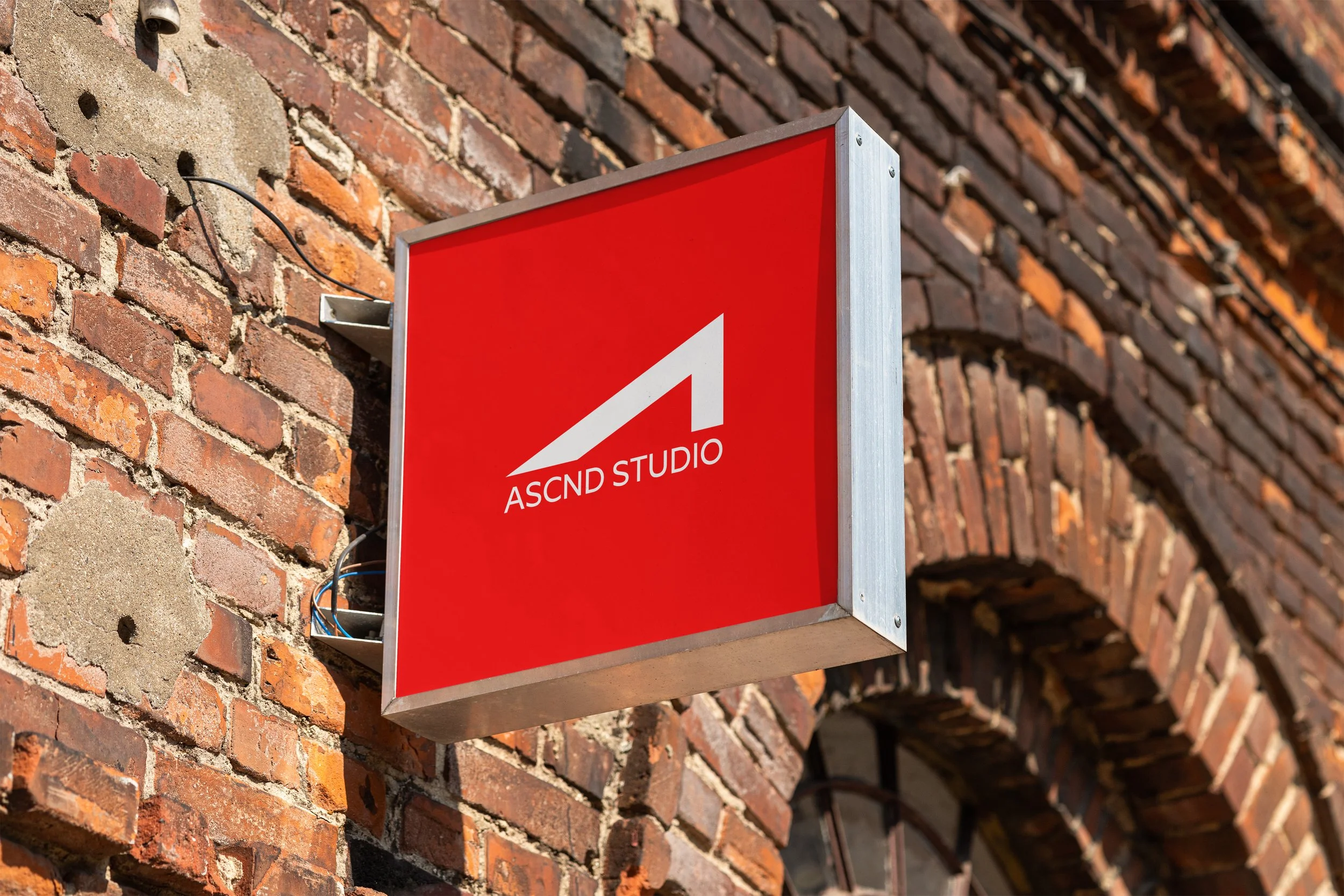 Red square sign with a white upward-sloping triangle and the text 'ASCND STUDIO' mounted on a brick wall.