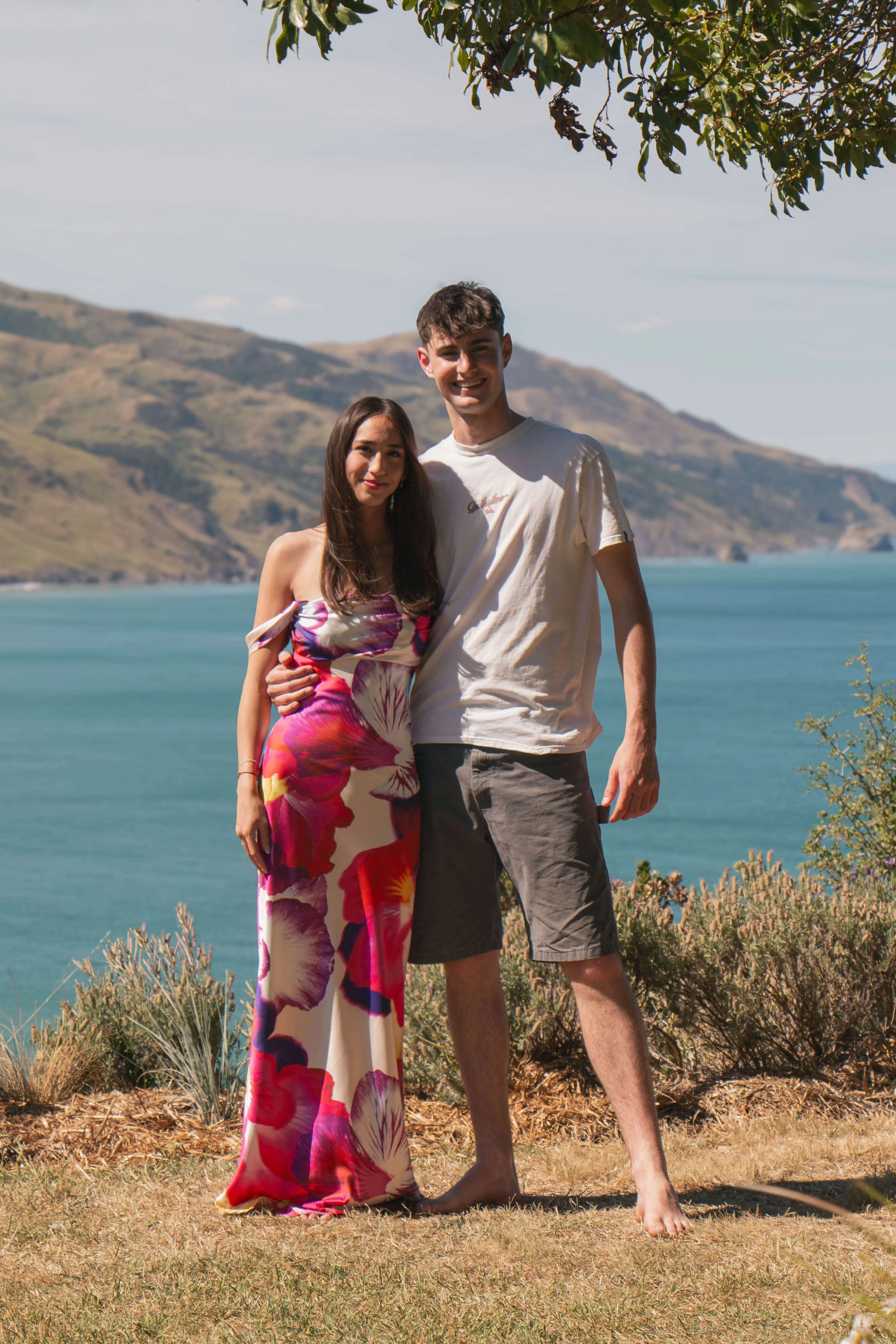 A young couple standing outdoors by a lake with hills in the background. The woman is wearing a colorful floral dress, and the man is wearing a white t-shirt and shorts. They are smiling and embracing each other.