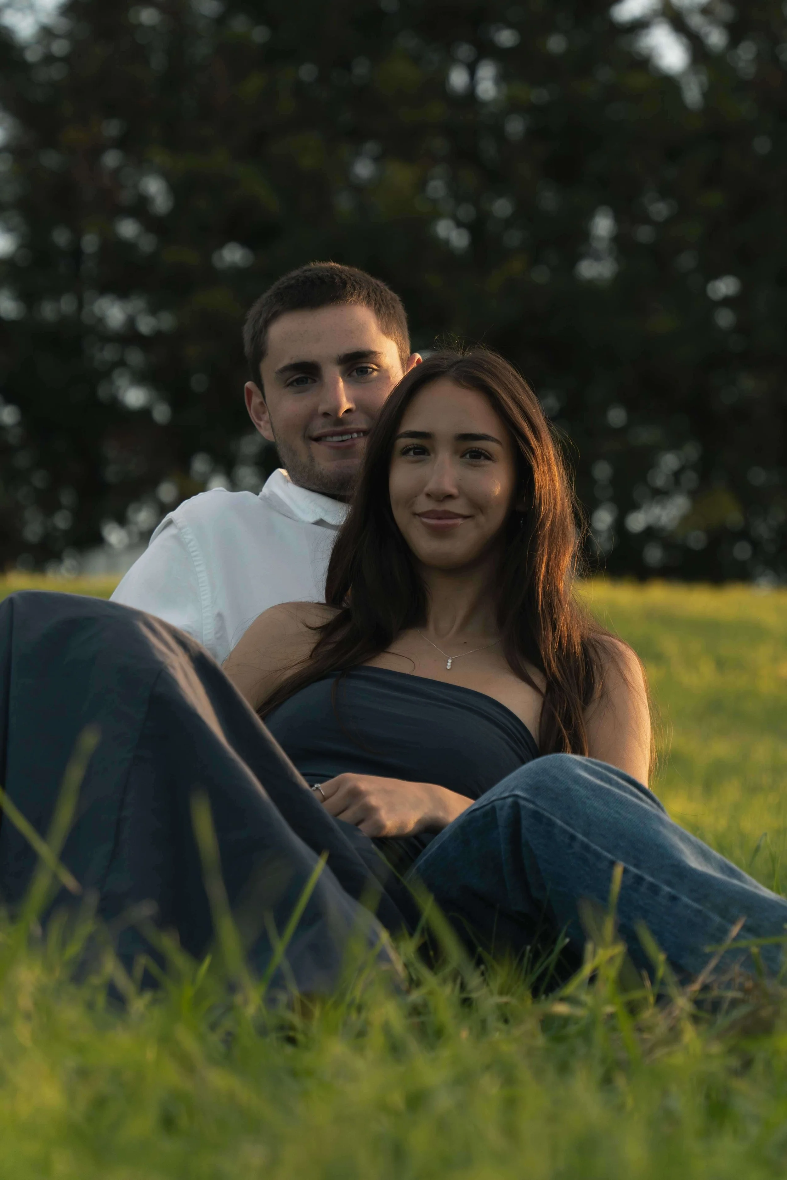 A young couple sitting outdoors on grass in evening light, with trees in the background.
