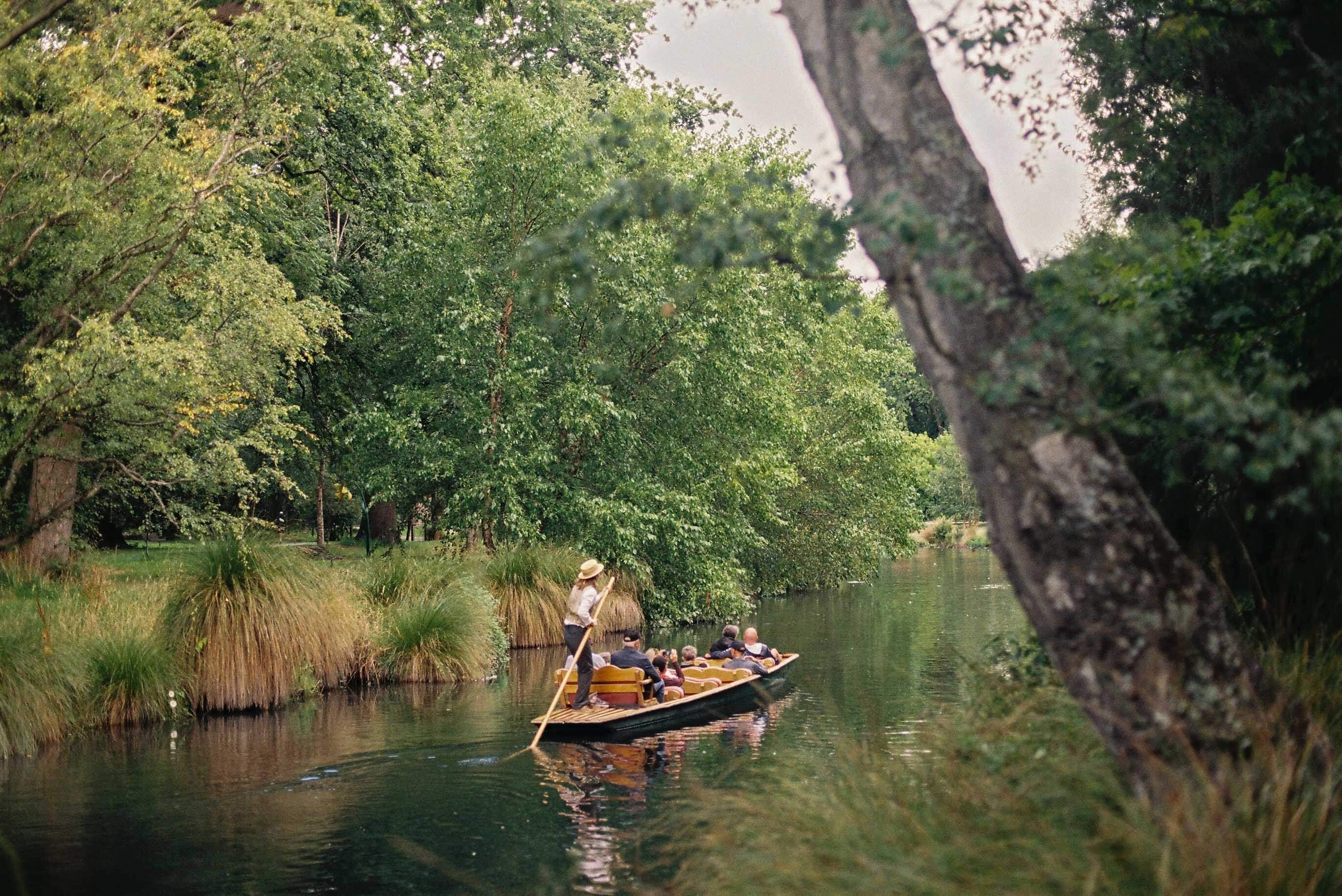 A group of people on a boat being guided through a lush, green river by a boat operator with a straw hat, surrounded by dense trees and grass