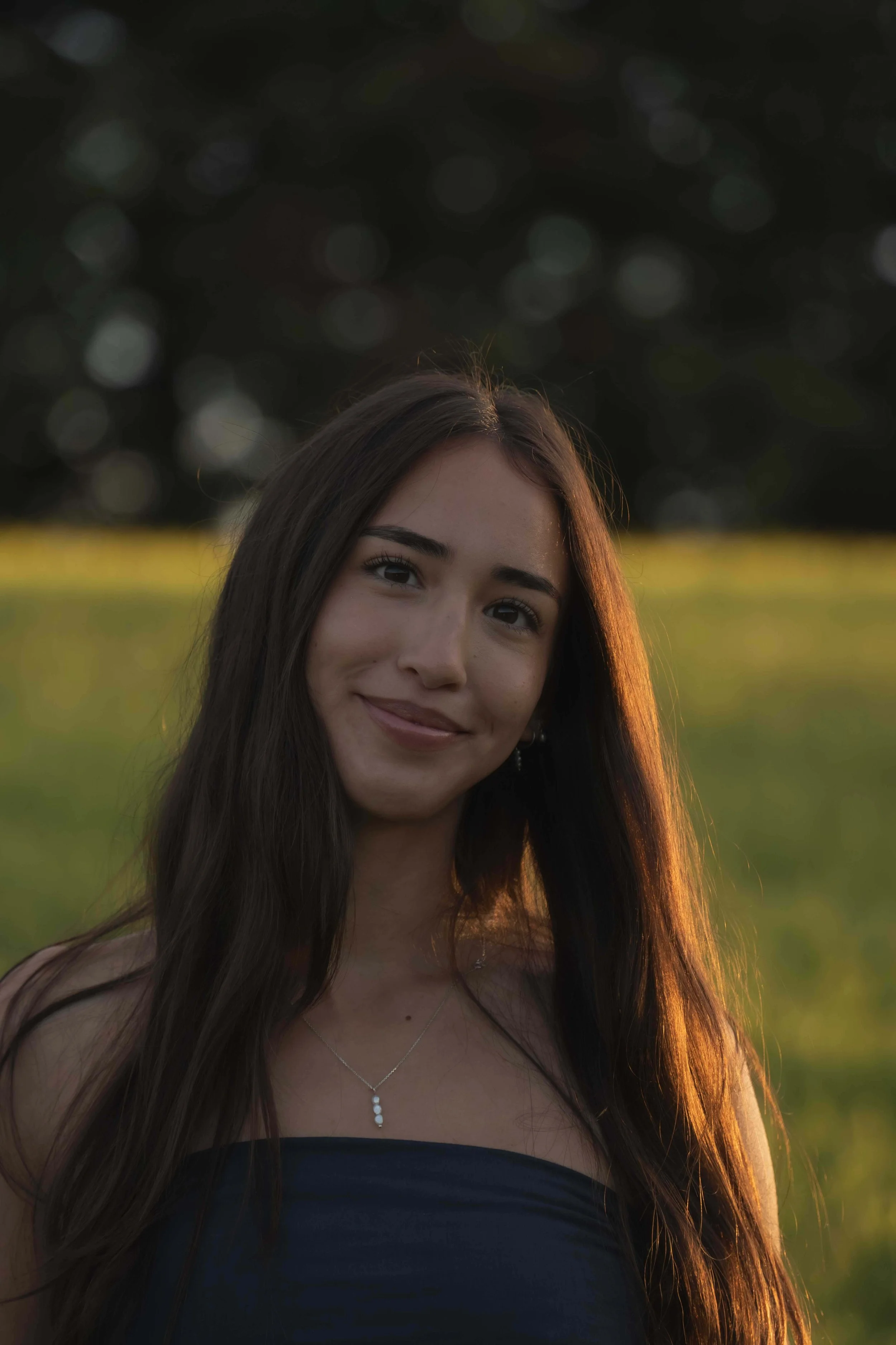 A young woman with long brown hair smiling outdoors during sunset, wearing a dark strapless top and silver jewelry.