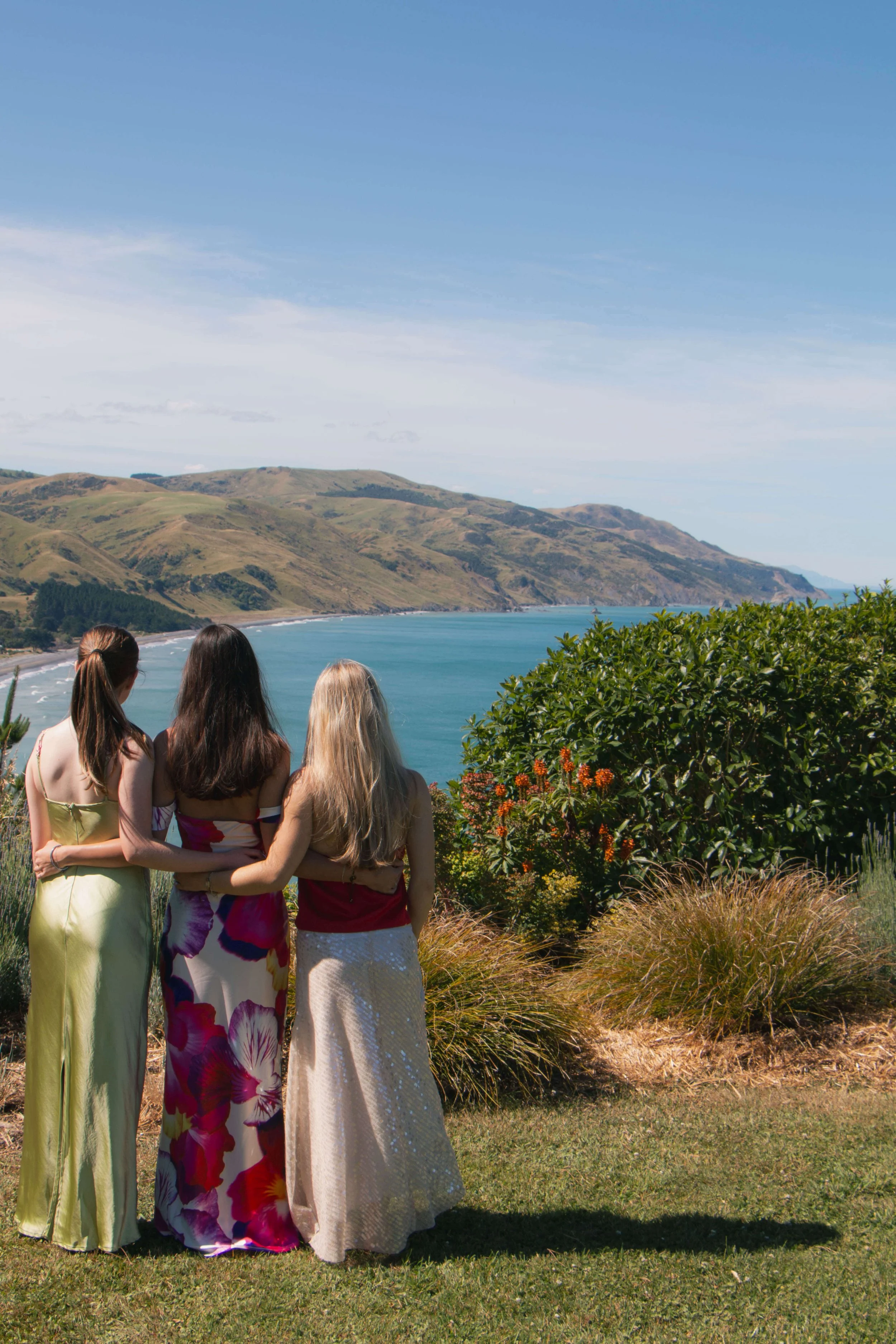 Three women in summer dresses standing arm-in-arm on a grassy hill overlooking a coastline with hills, water, and a partly cloudy sky.