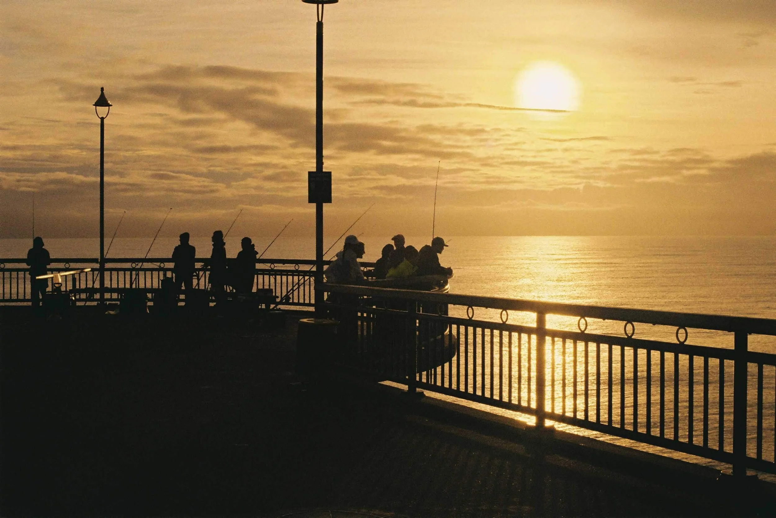 Silhouettes of people fishing along a seaside promenade at sunset, with the ocean and a golden sky in the background.