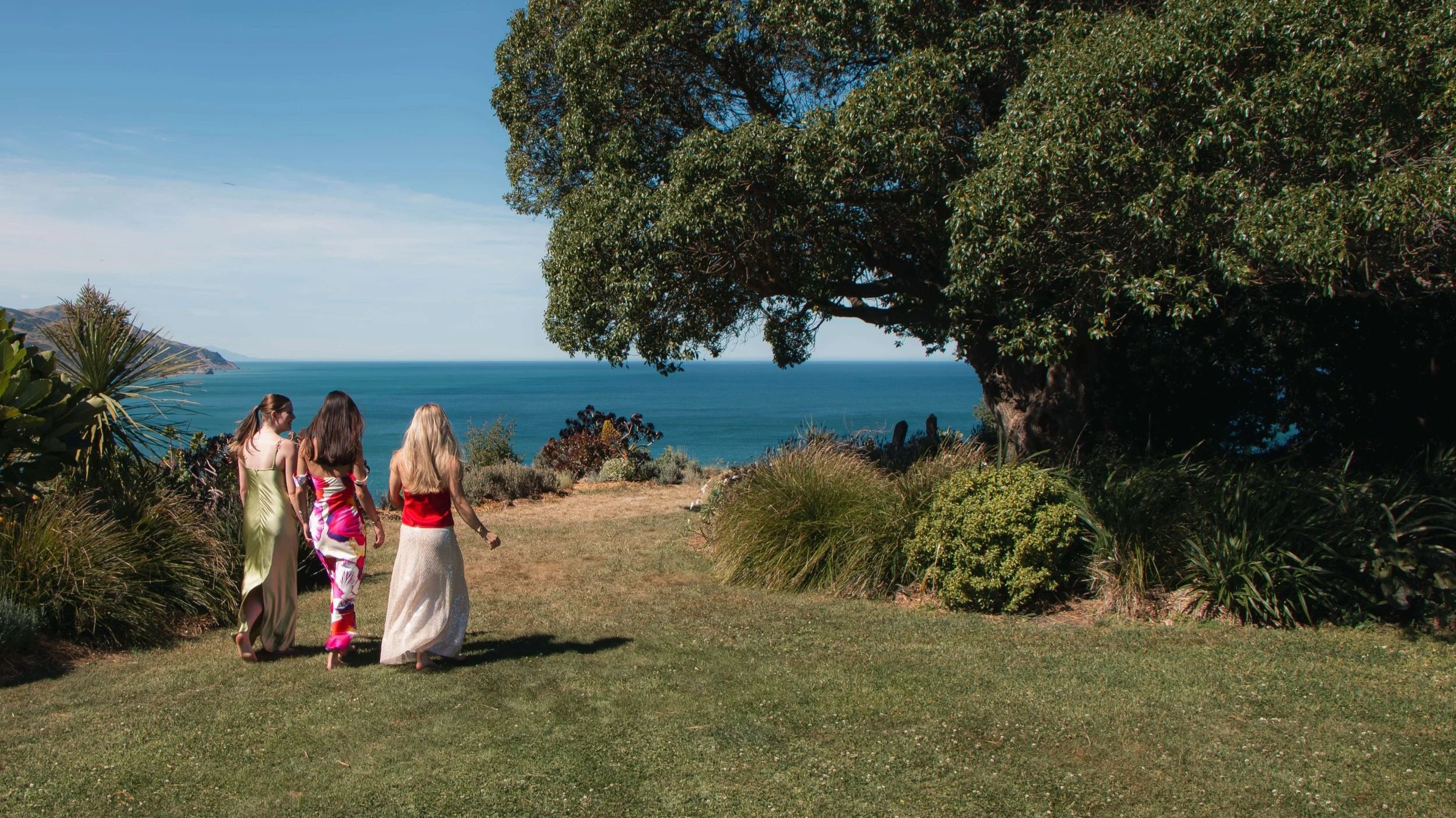Three women in colorful dresses walking on a grassy path near a large tree, overlooking the ocean with a coastline in the distance.