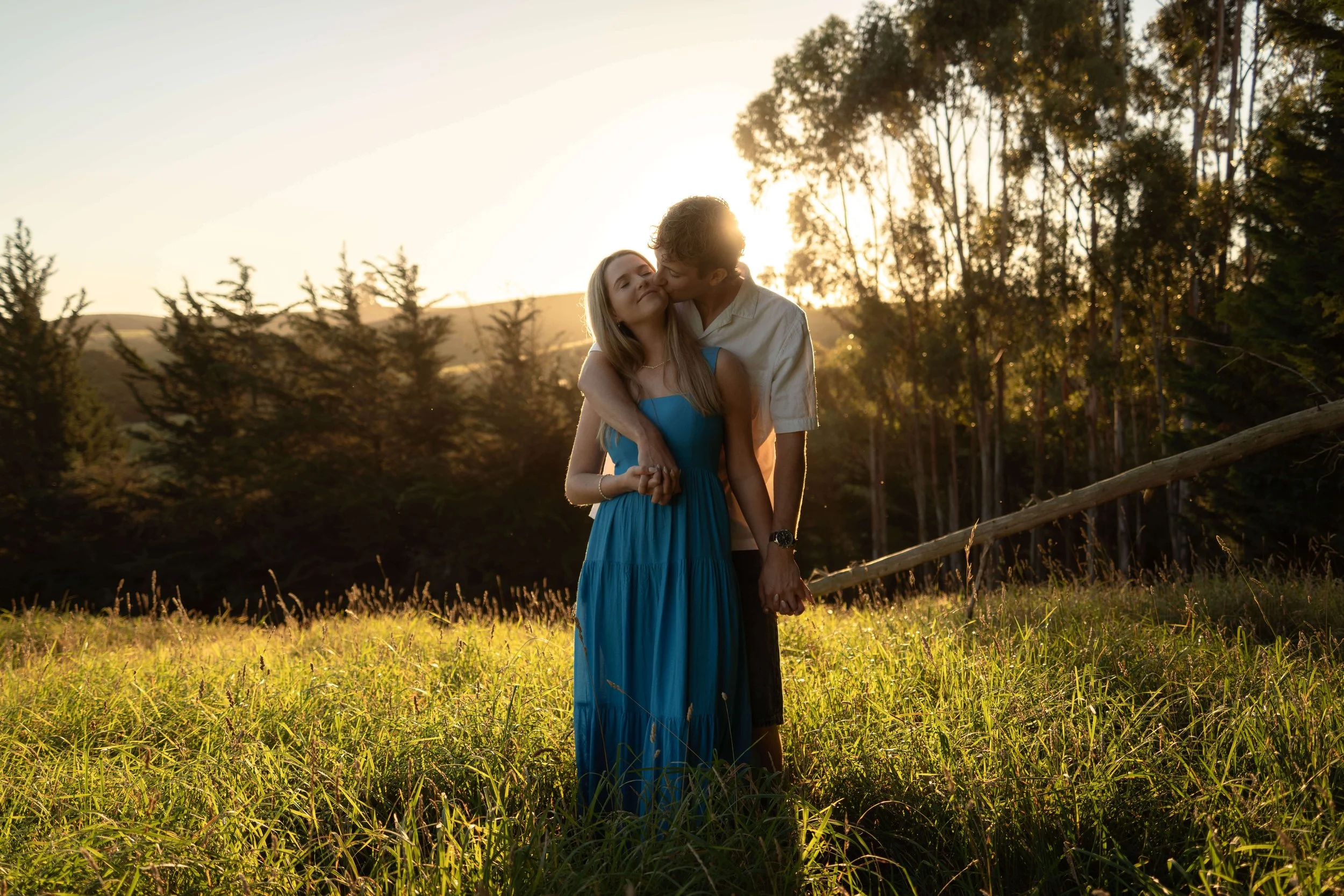 A couple holding hands and sharing a kiss in a grassy field during sunset, with trees and hills in the background.