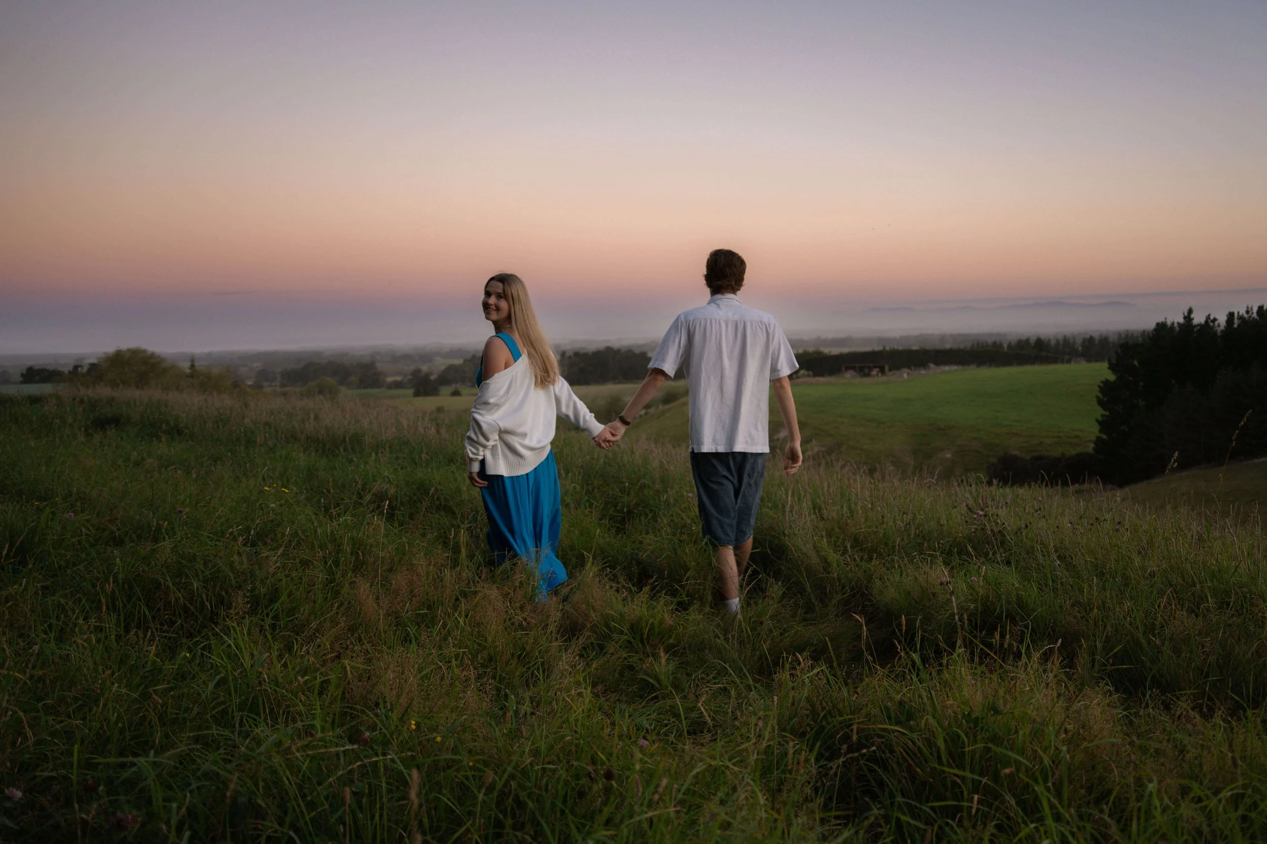 A couple holding hands while walking in a grassy field during sunset, with a colorful sky and distant trees.