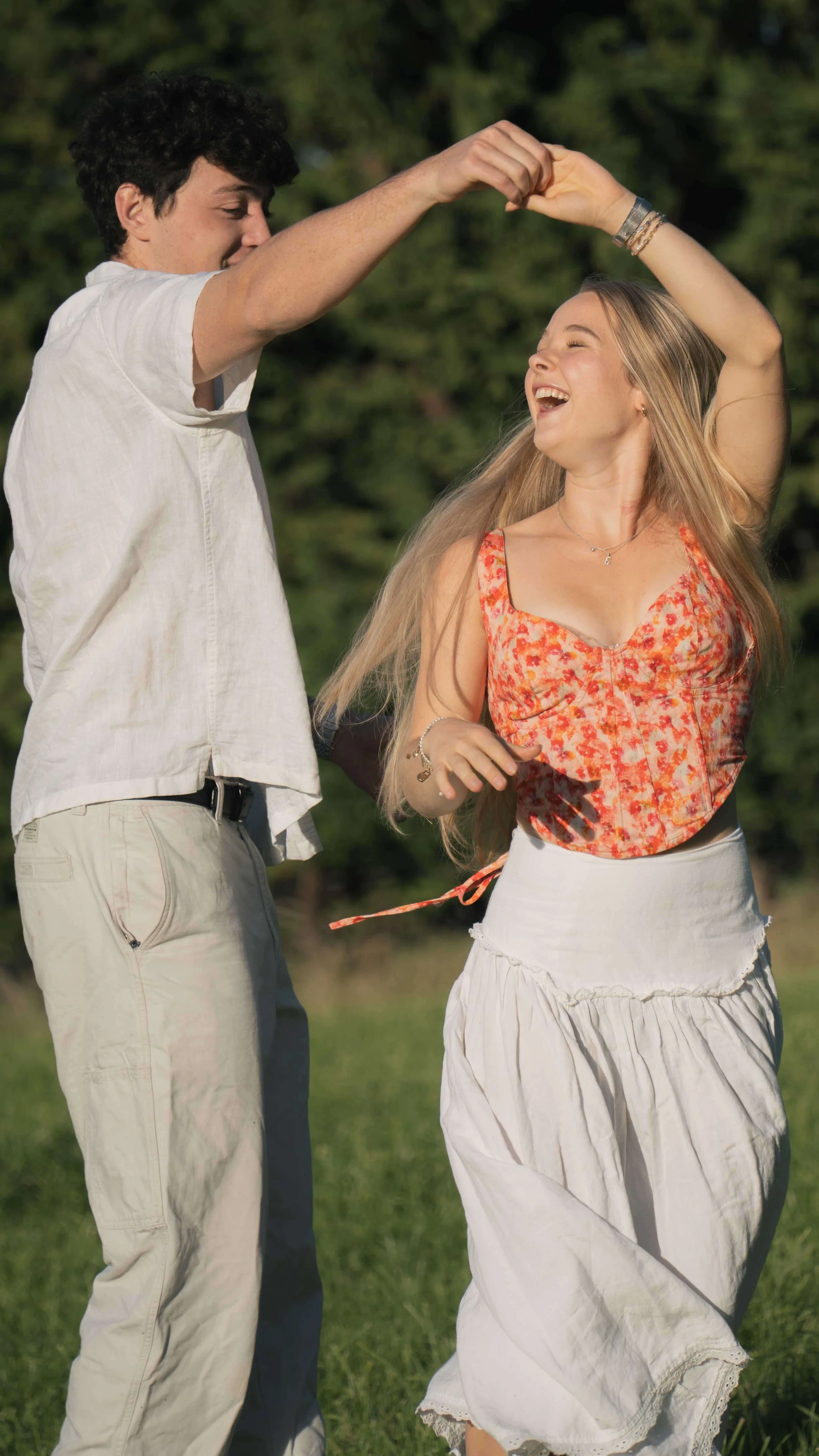 A young man and woman are dancing or spinning outdoors in a grassy area with trees in the background. They are smiling and appear to be enjoying themselves.