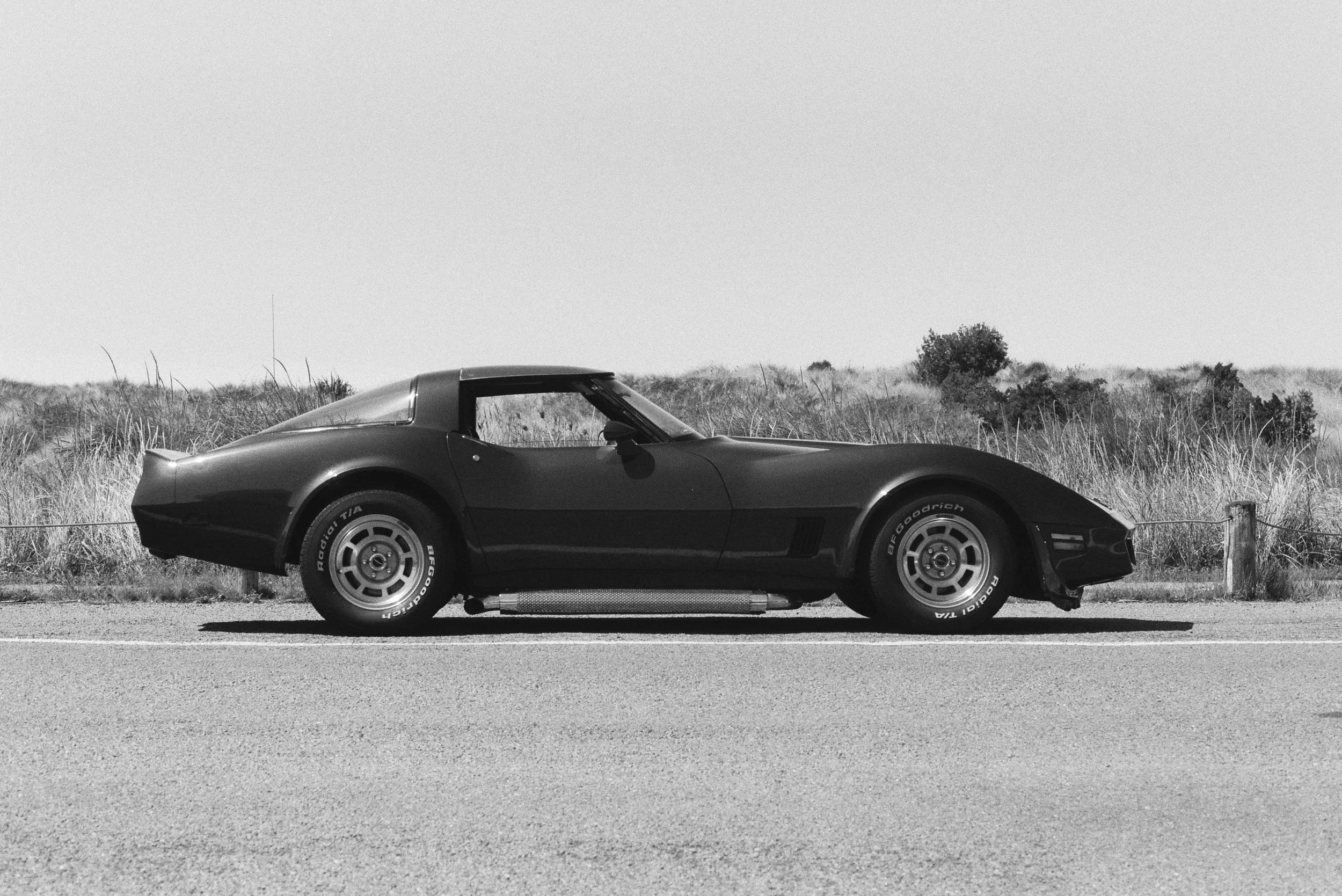 A vintage black sports car parked on a road beside a grassy field with some bushes, in a black and white photo.