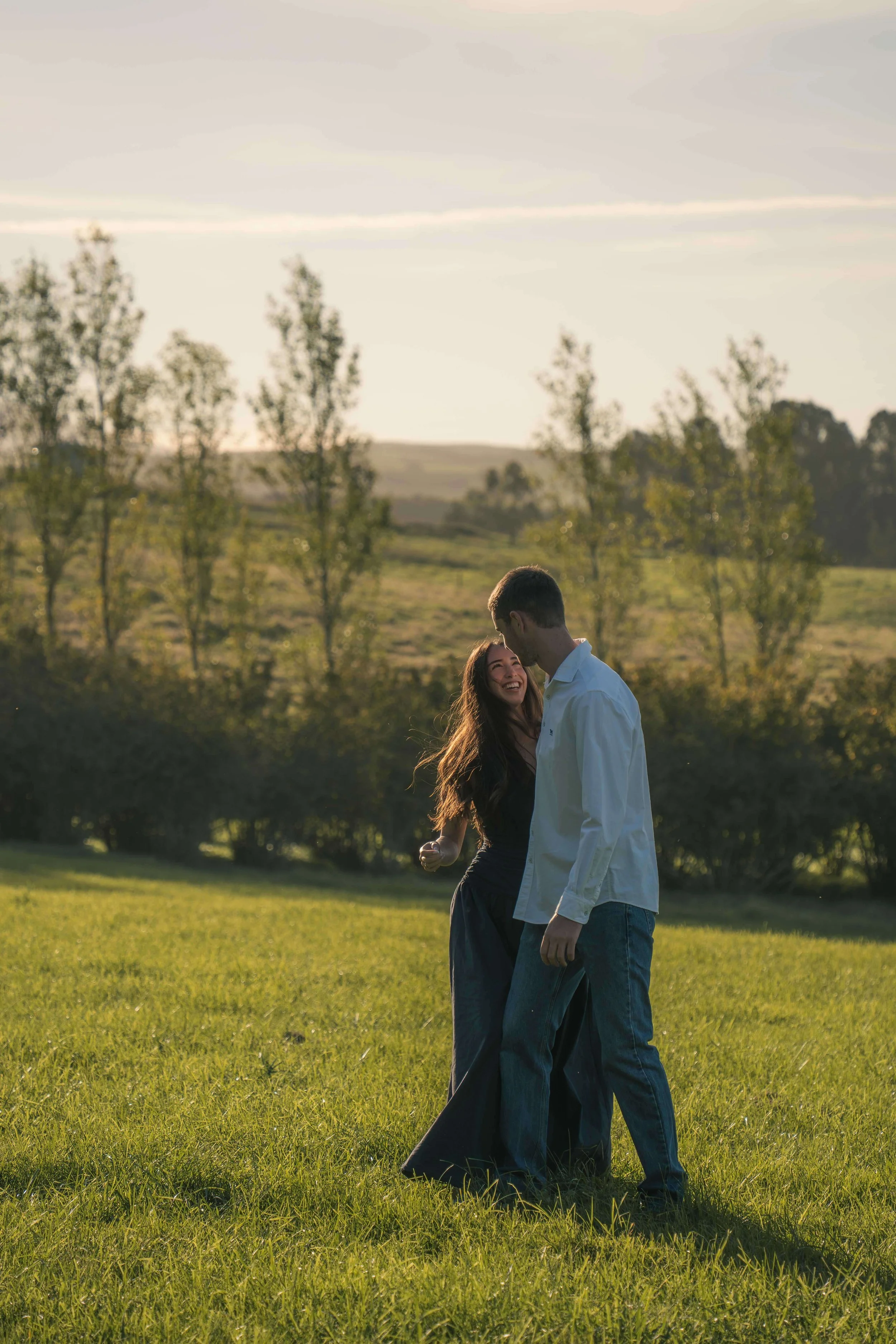 A couple is standing on a grassy field, smiling and enjoying each other's company with a backdrop of trees and distant hills during sunset.