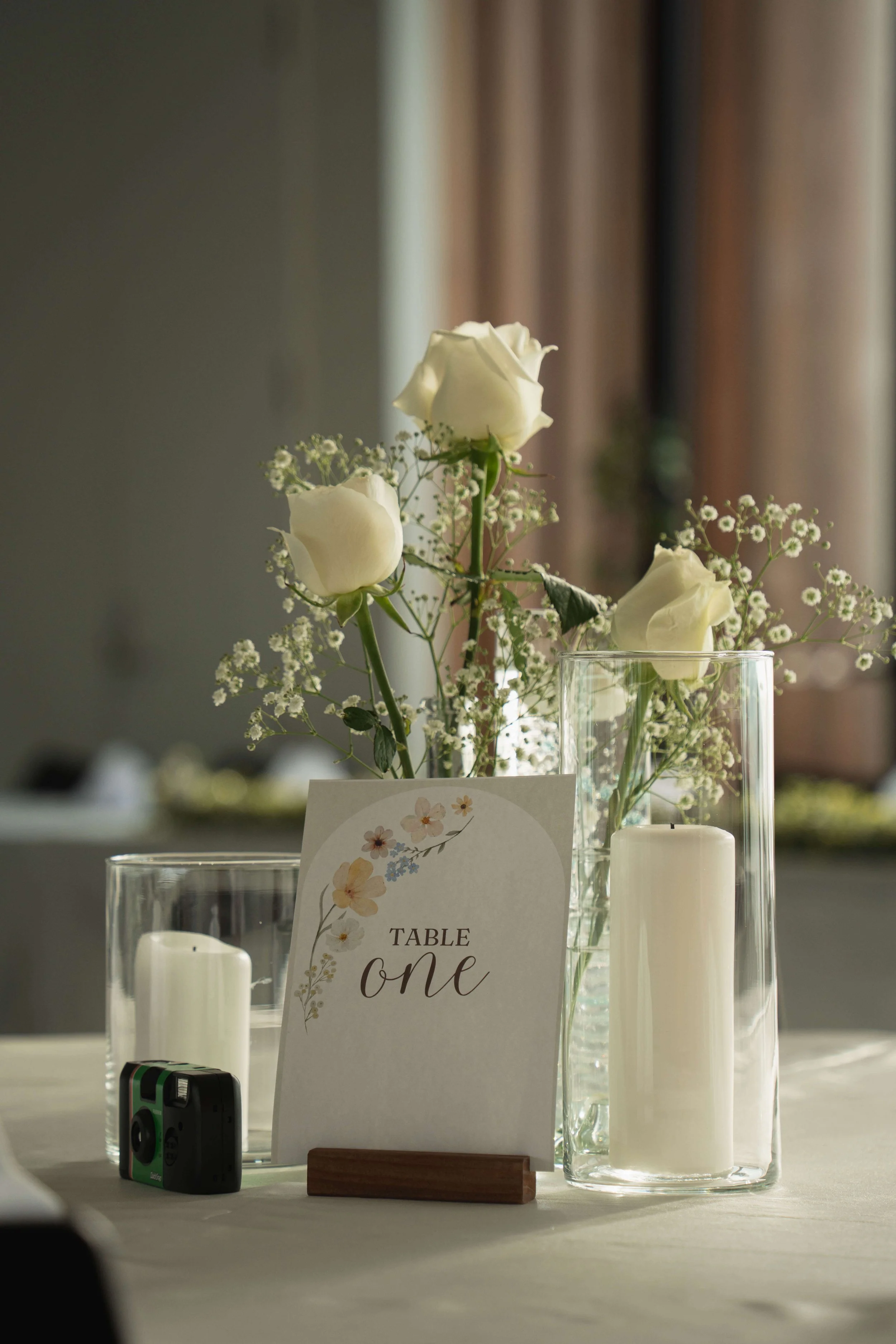 A table centerpiece with white roses, baby's breath, and candles, along with a sign reading 'Table One' at an event.