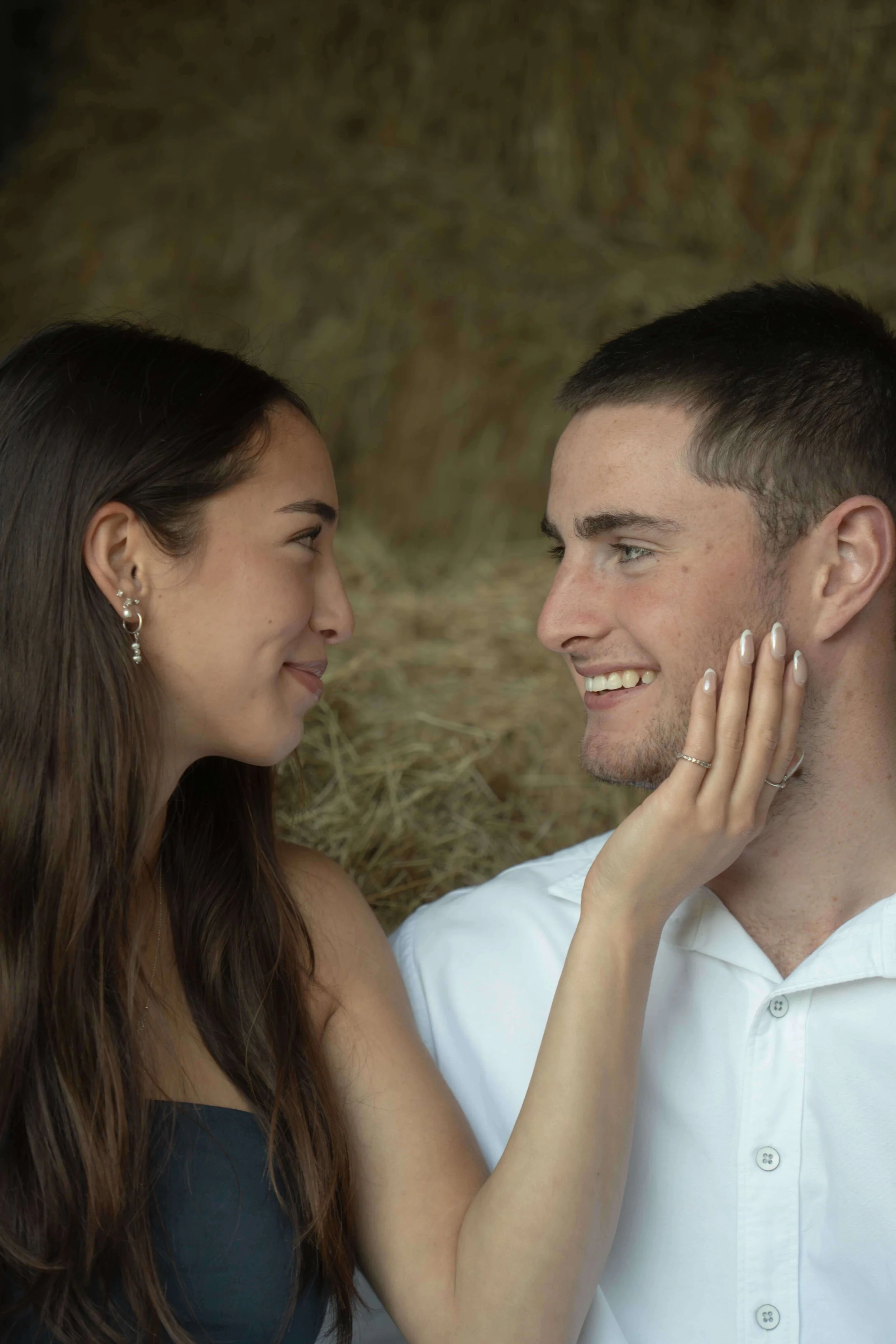 A woman gently touching a smiling man's face, both looking into each other's eyes, with a blurred natural background.