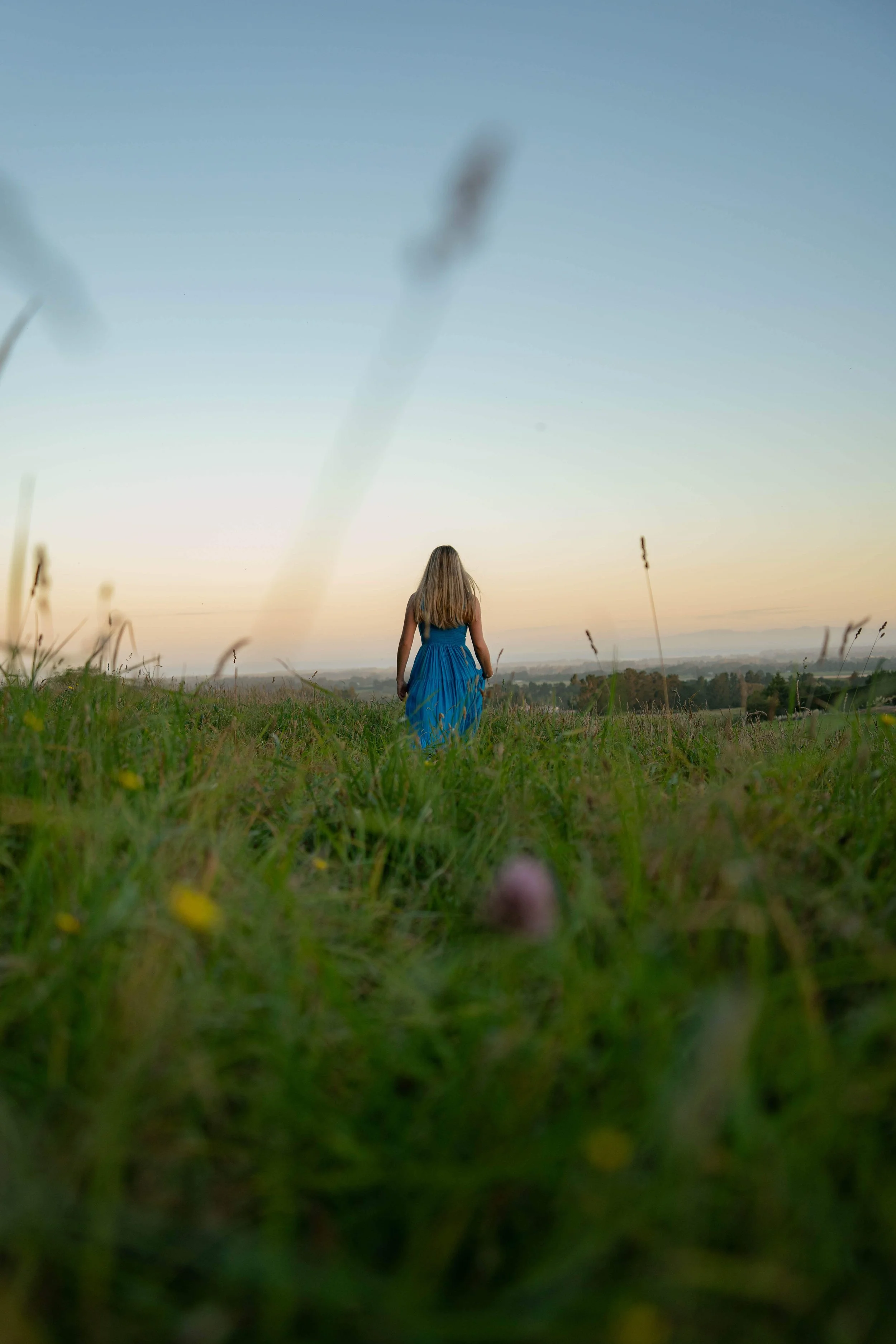 A woman in a blue dress with long blonde hair walking through a grassy field at sunset, with a distant landscape and clear sky in the background.