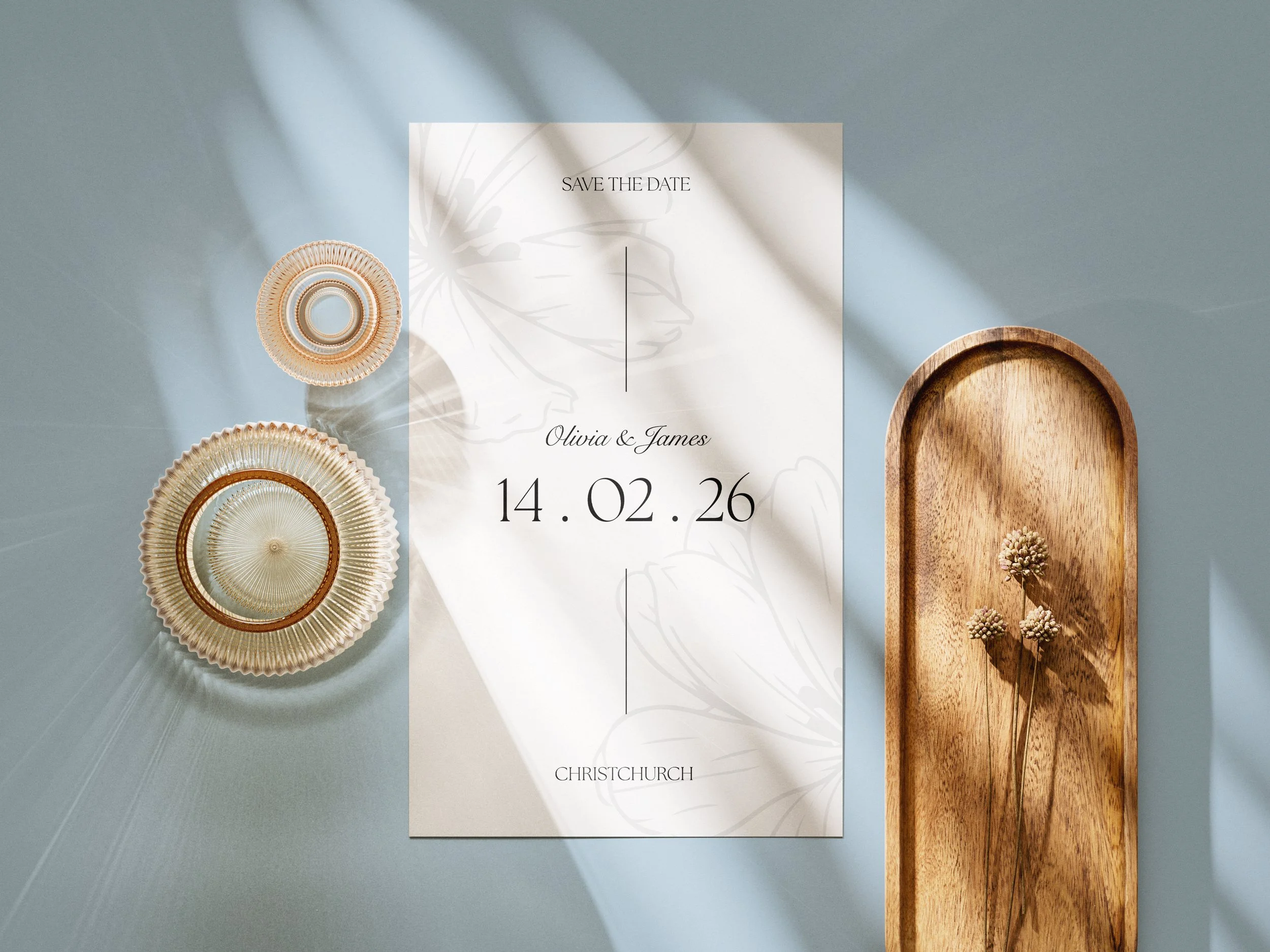 A wedding save the date card for Olivia and James with the date 14.02.26, placed on a light blue surface. To the left are two decorative paper circles, and to the right is a wooden tray with small dried flowers inside. Shadows from sunlight are cast across the items.