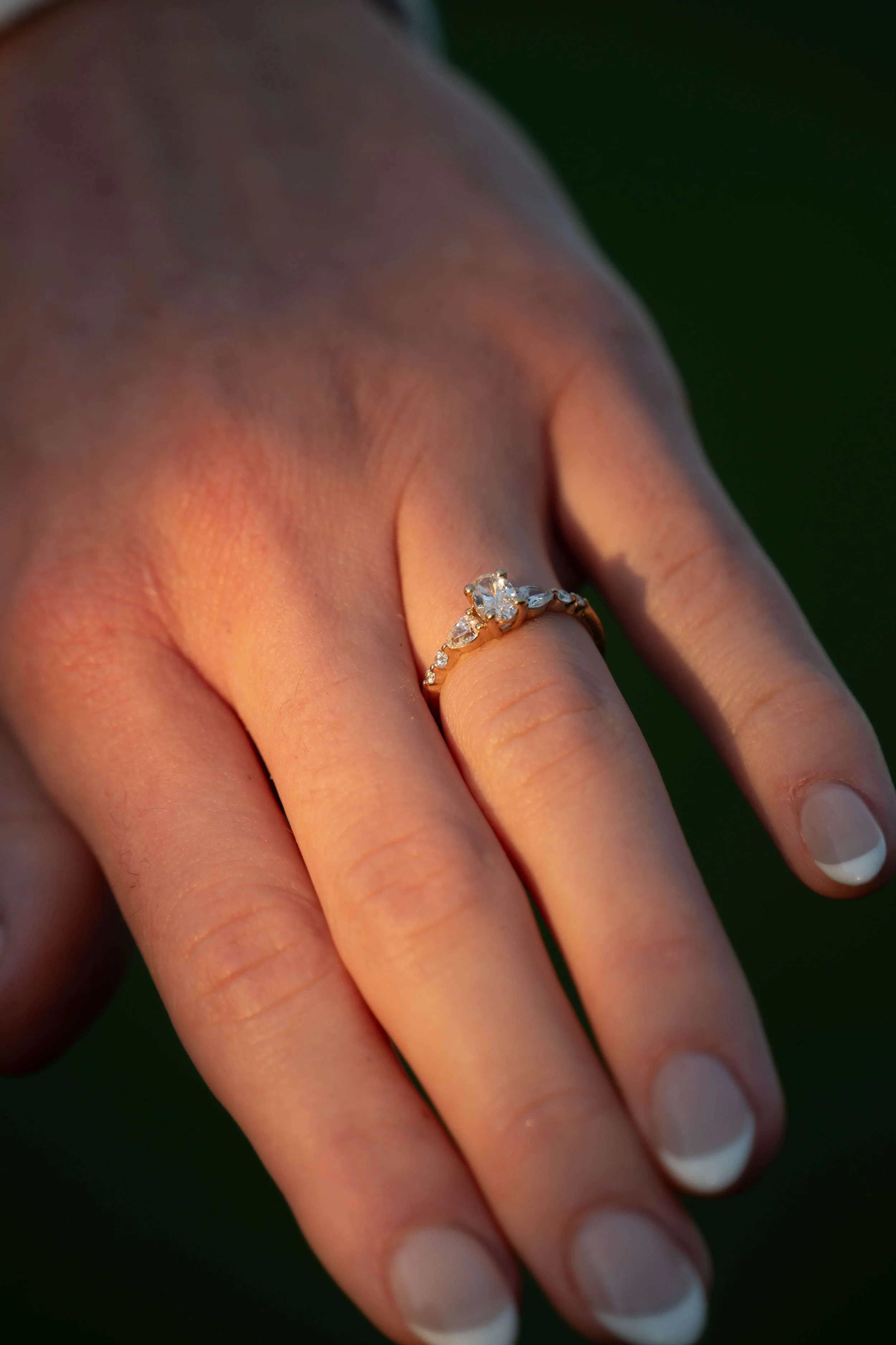 Close-up of a woman's hand showing an engagement ring with a large central diamond, surrounded by smaller diamonds on a gold band, against a dark background.