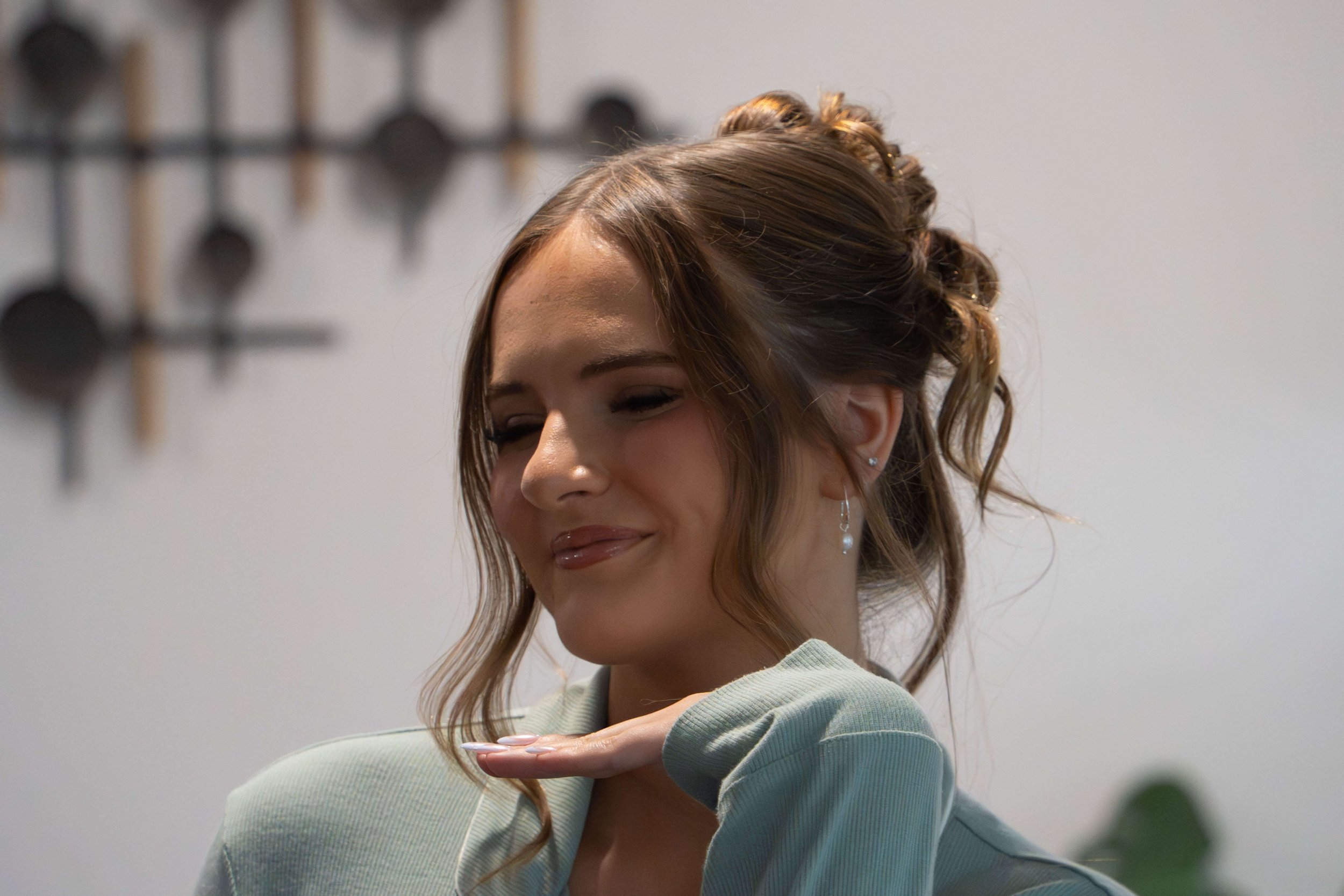 A woman with long, wavy brown hair styled in an elegant updo, wearing earrings, with her eyes closed and a gentle smile, resting her chin on her hand.