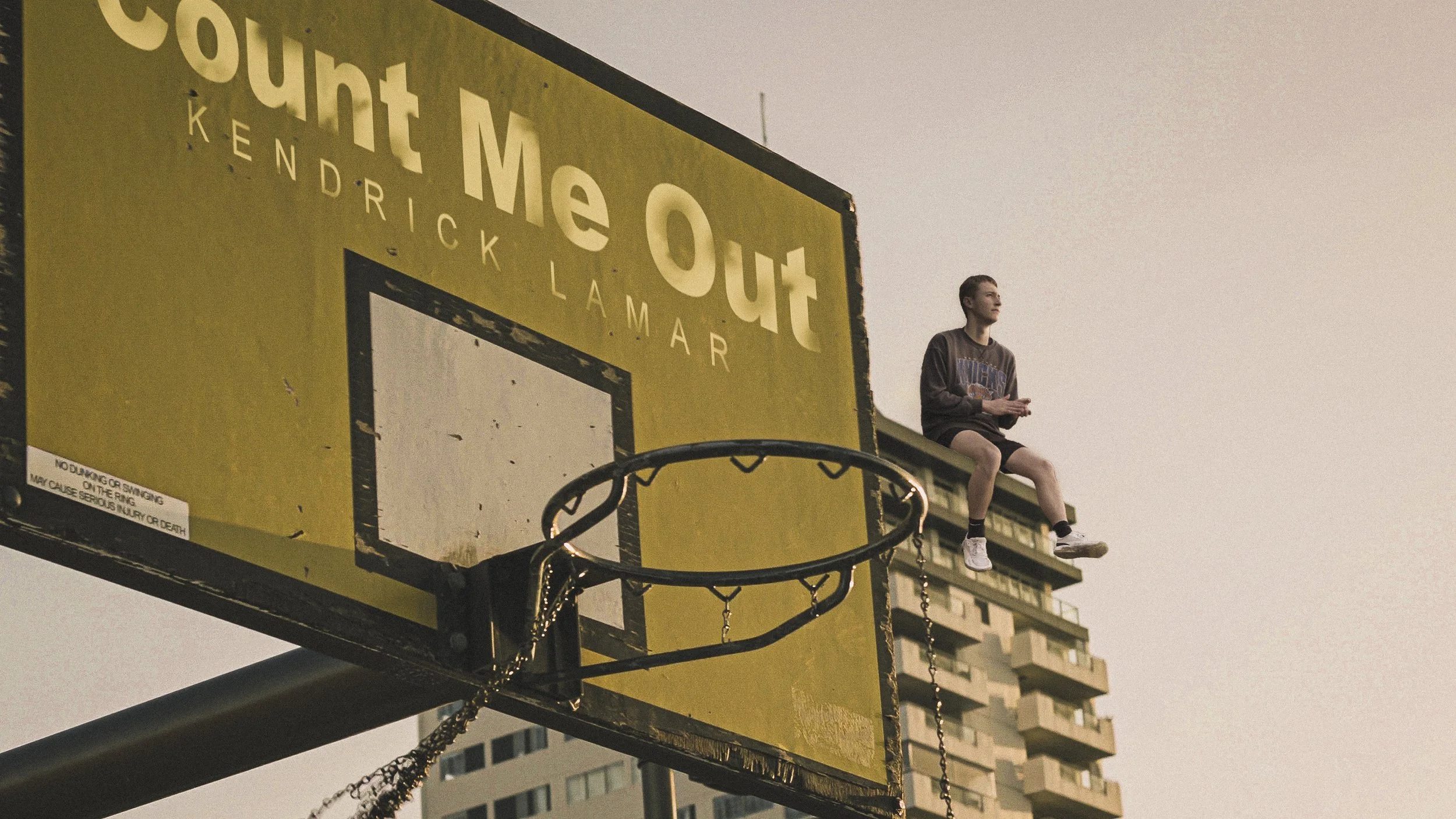 A person sitting on the edge of a basketball backboard during sunset. The backboard has a yellow advertisement with the text 'Count Me Out' and 'Kendrick LaMar'. The setting appears urban, with tall buildings in the background.