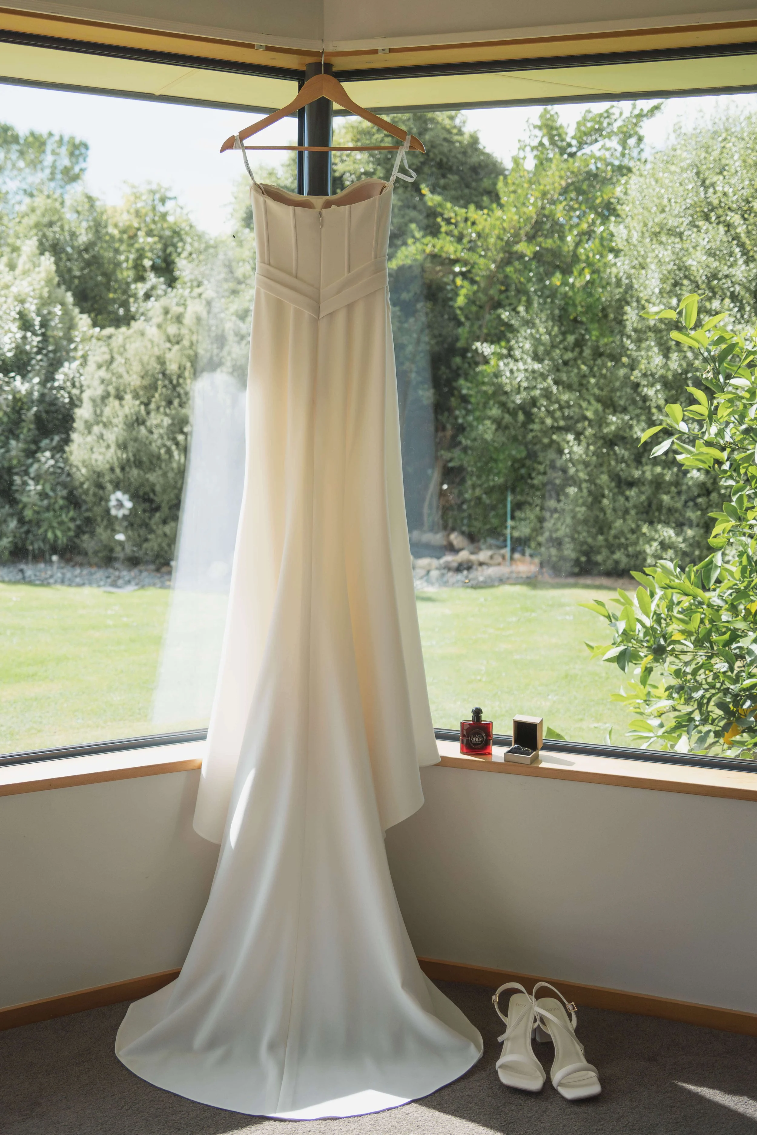 A white wedding dress hanging on a wooden hanger in front of a large window, with a pair of white heels on the floor beneath the dress, and perfume bottles on the windowsill.