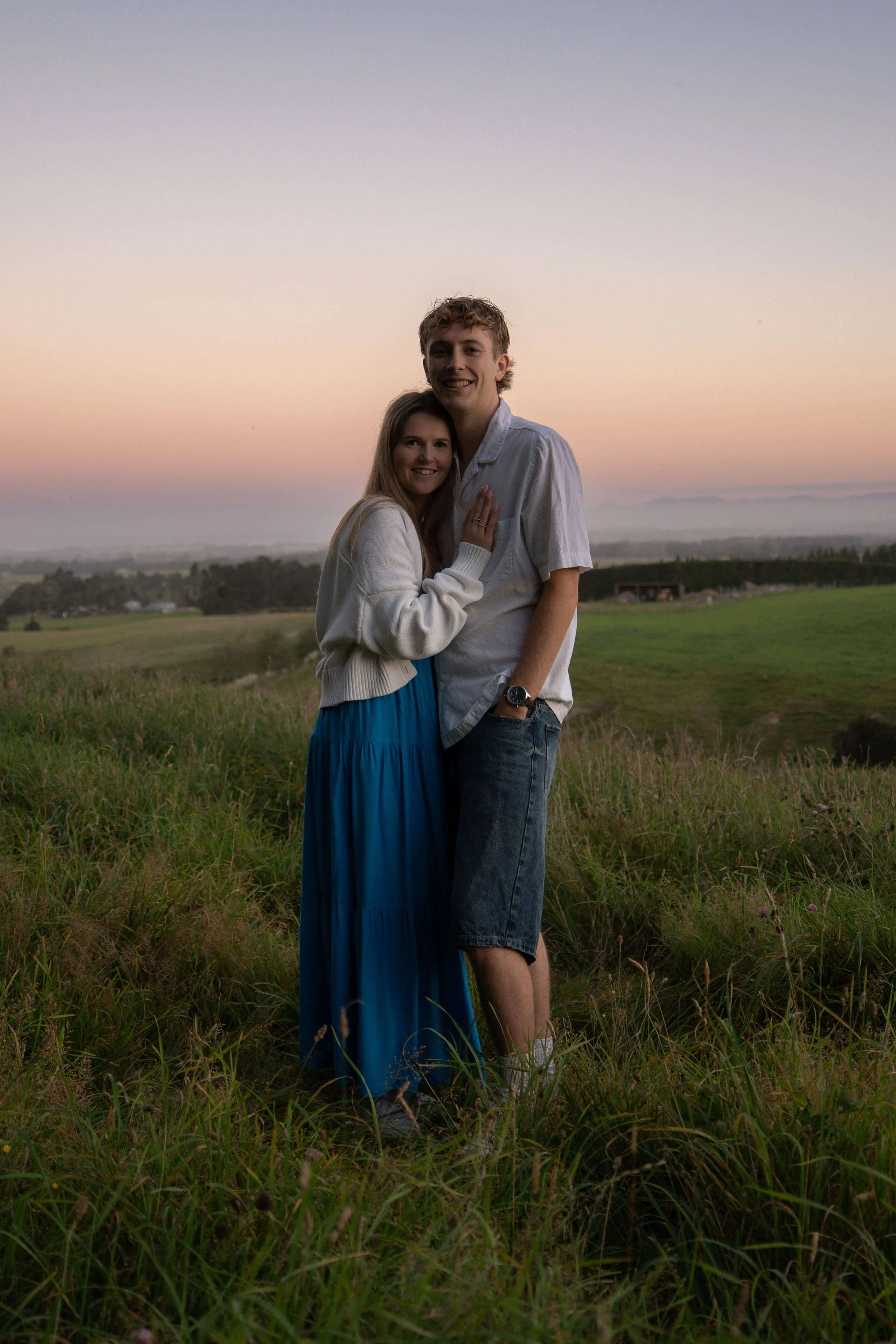 A young couple standing together outdoors during sunset, smiling and hugging on a grassy field.