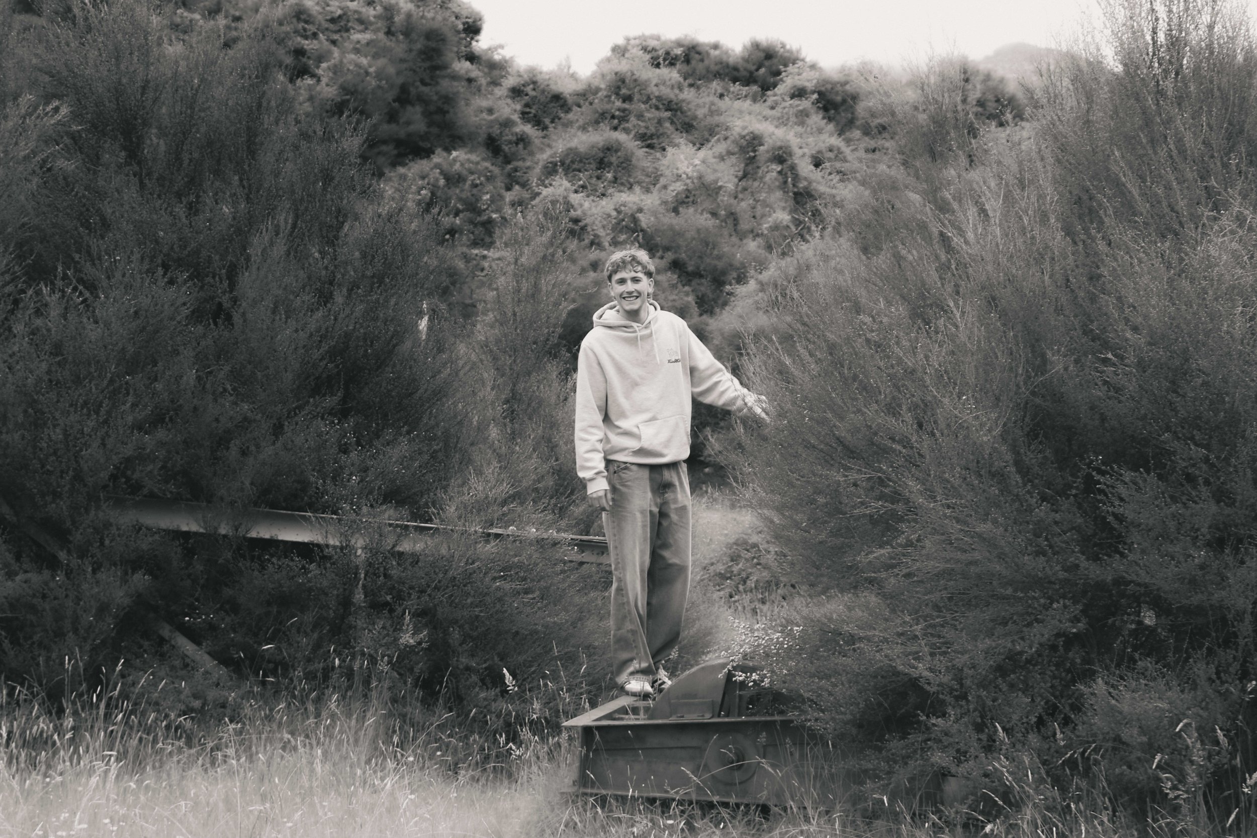 A young man standing  in a field surrounded by tall bushes and trees, smiling and wearing casual clothes.