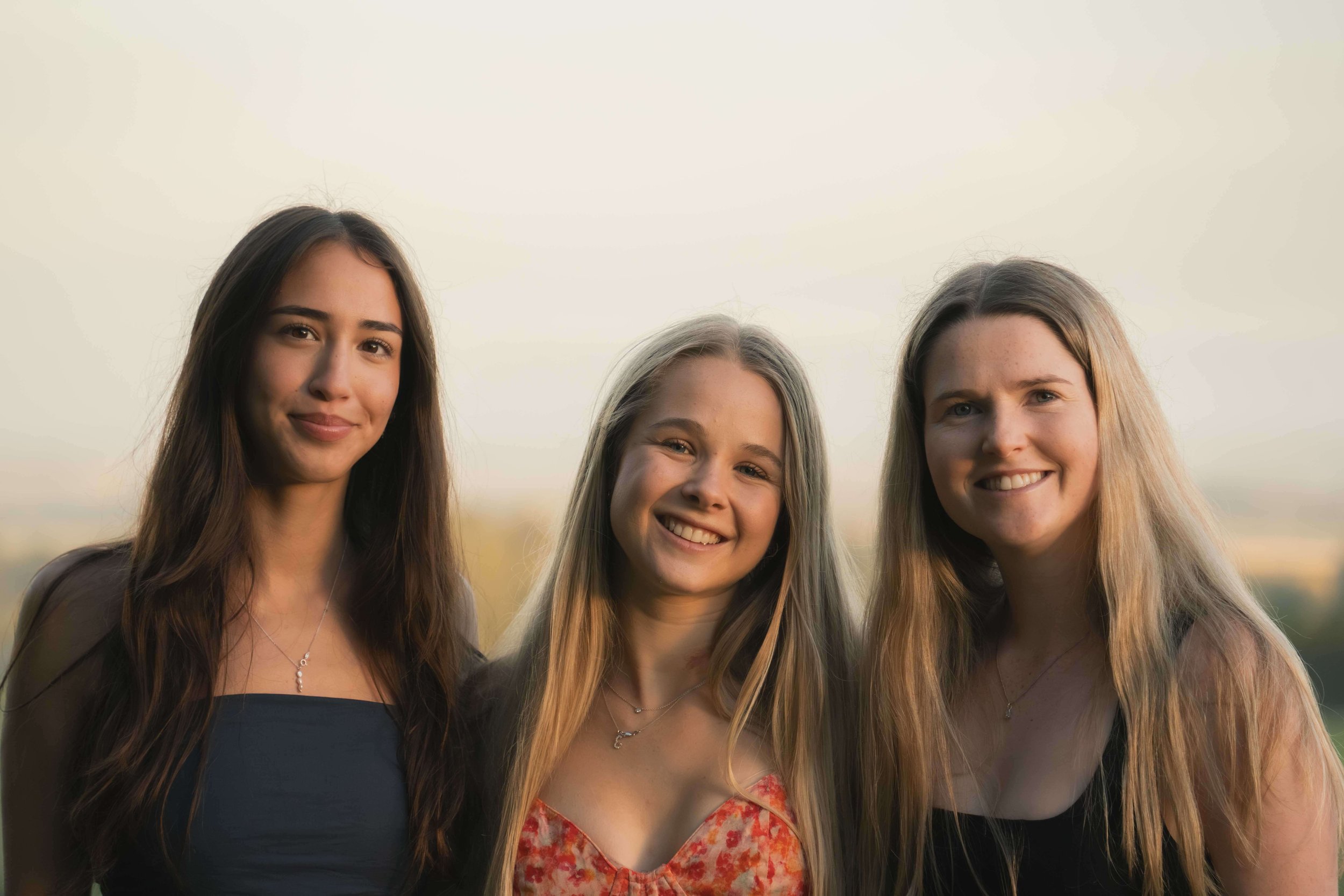 Three young women smiling outdoors with a blurred sky background.