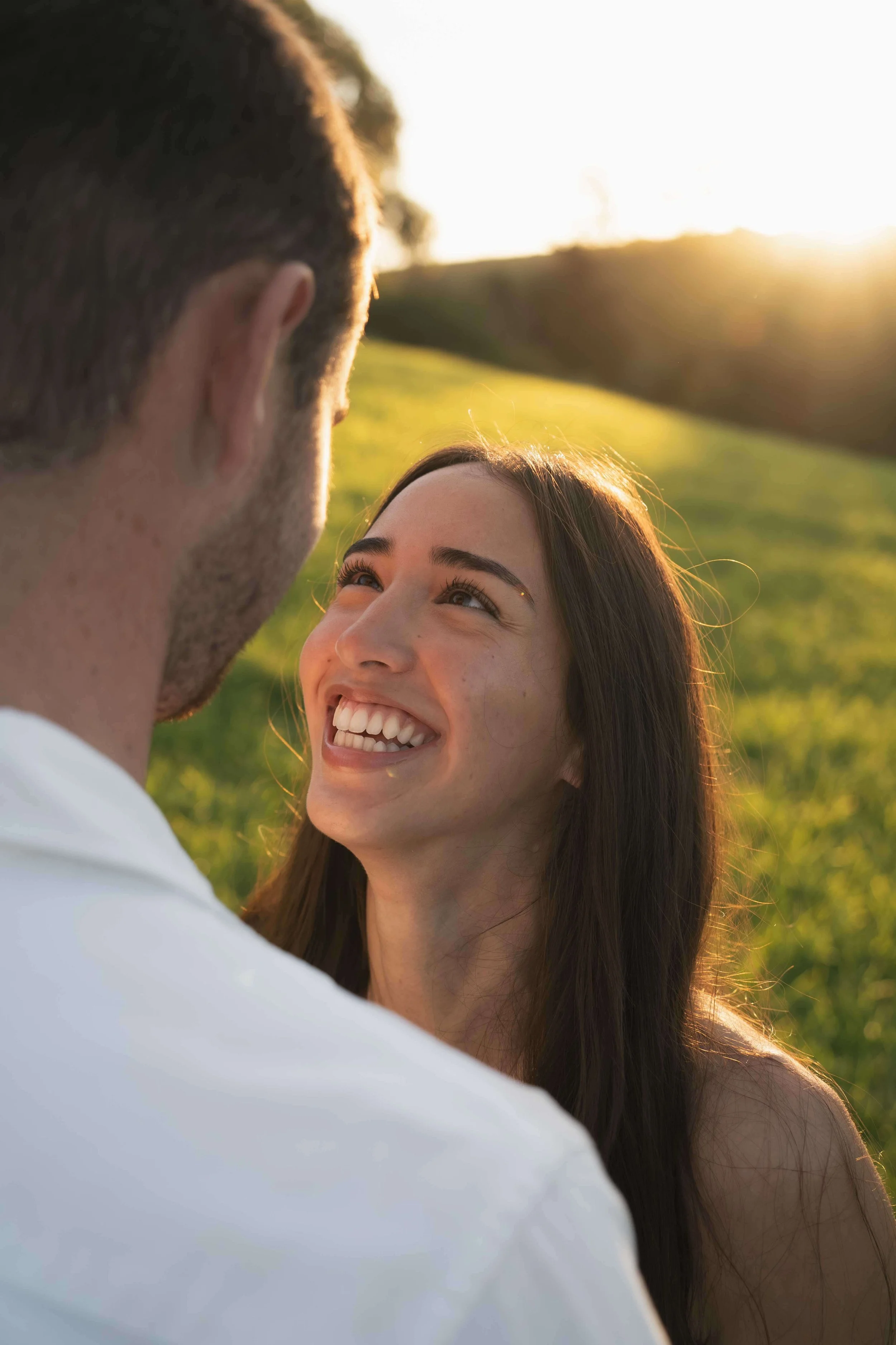 A woman smiling and looking at a man during sunset outdoors, with green grass and hills in the background.