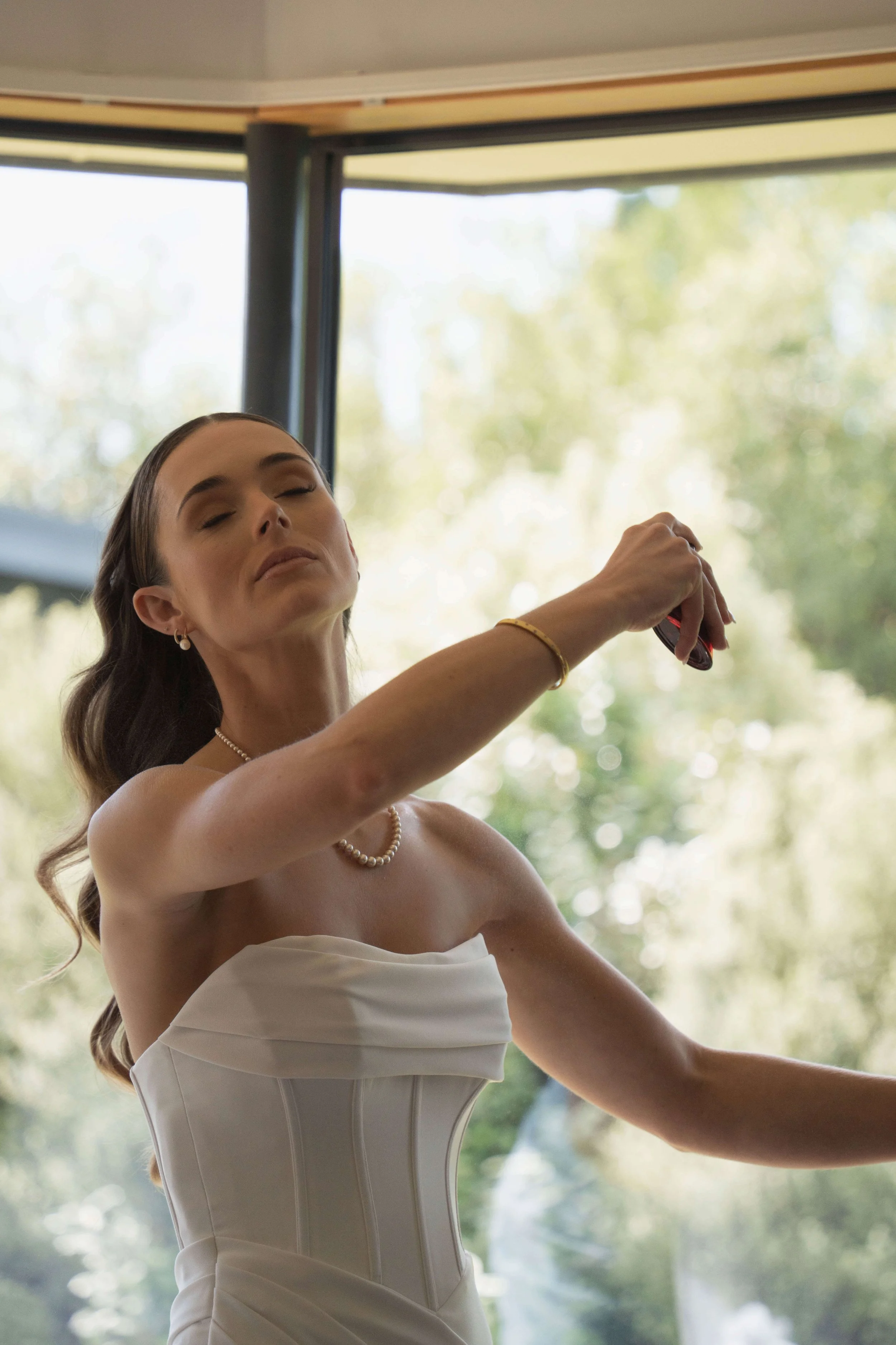 A woman wearing a white strapless dress and pearl necklace, with her eyes closed, appears to be relaxed or in a moment of calm, holding an object in her raised hand, against a background of large windows with a view of trees outside.