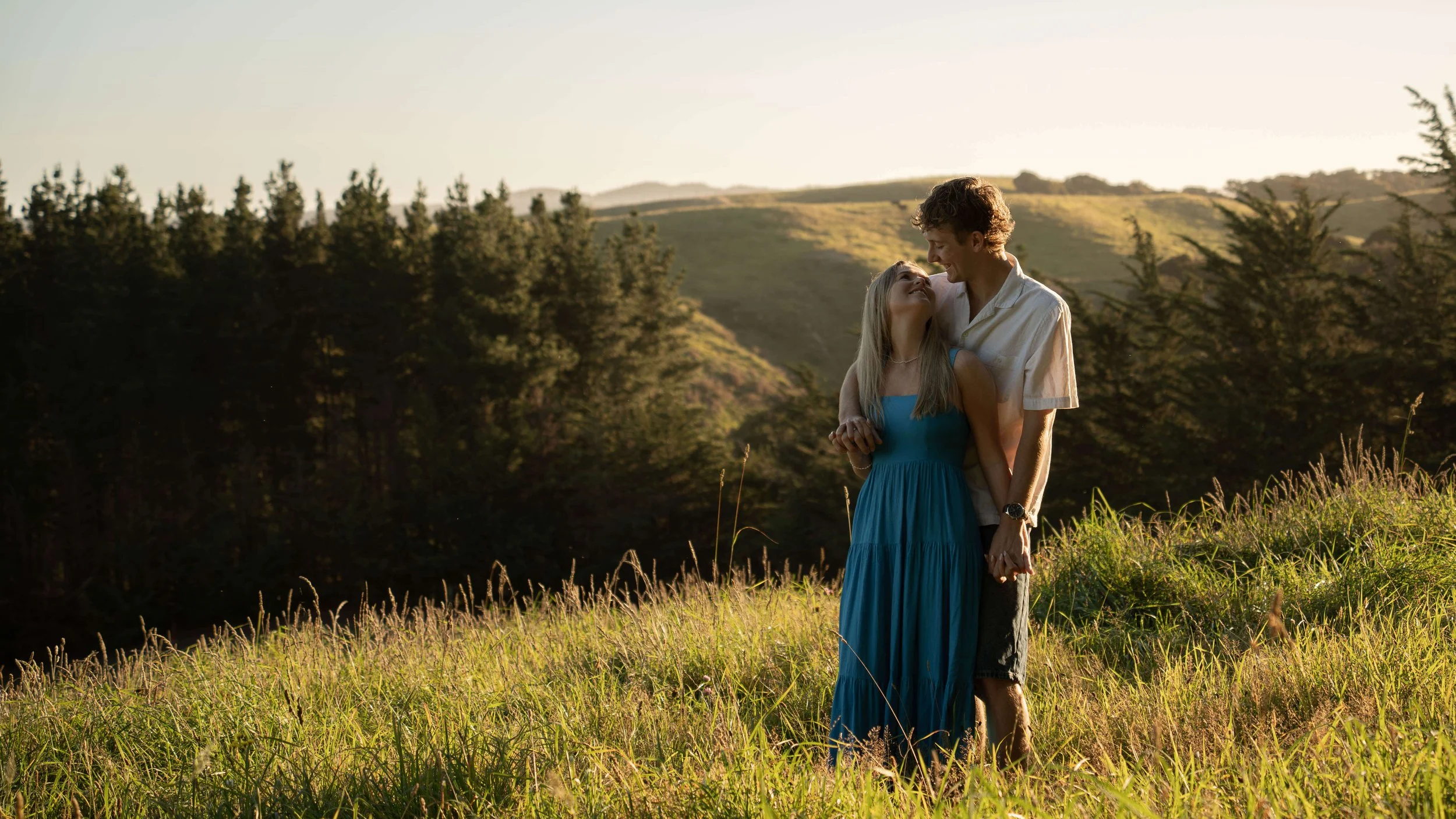A young couple standing in a field of tall grass, holding hands and gazing lovingly at each other during sunset.