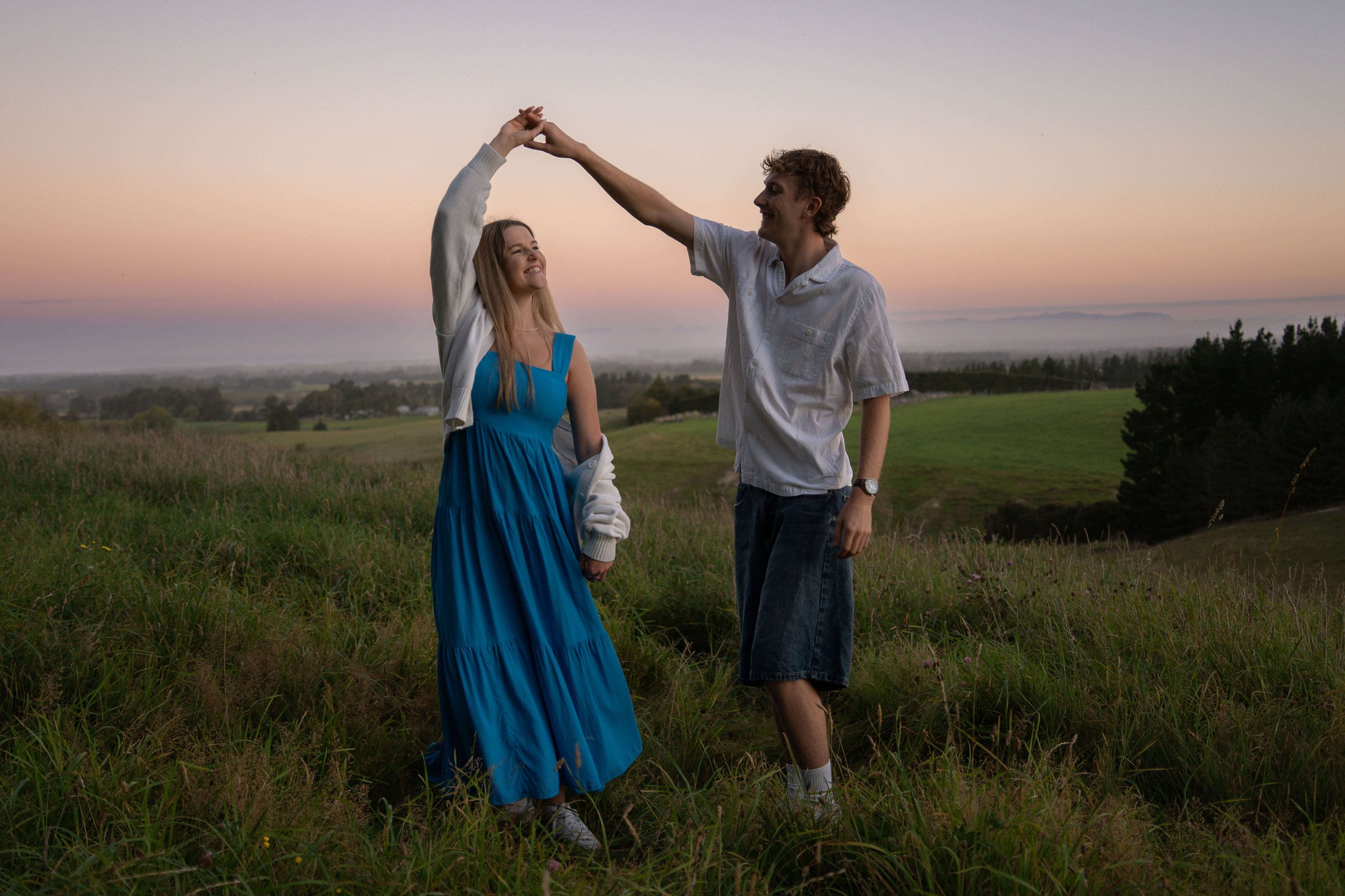 A young couple dancing in a grassy field during sunset with a scenic landscape in the background.