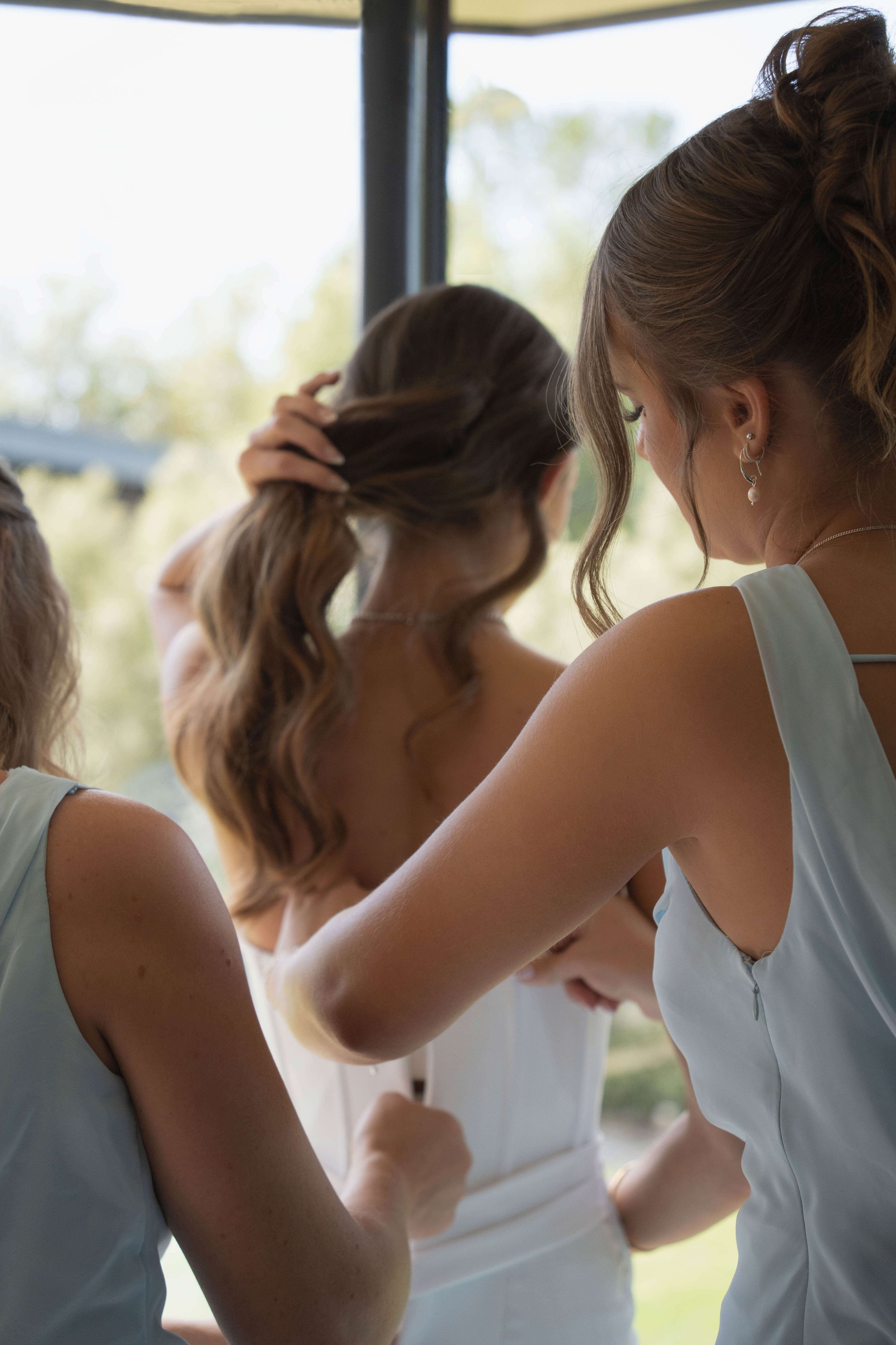 Three women in white dresses, possibly at a wedding, standing near a window with greenery outside, with one adjusting her hair.