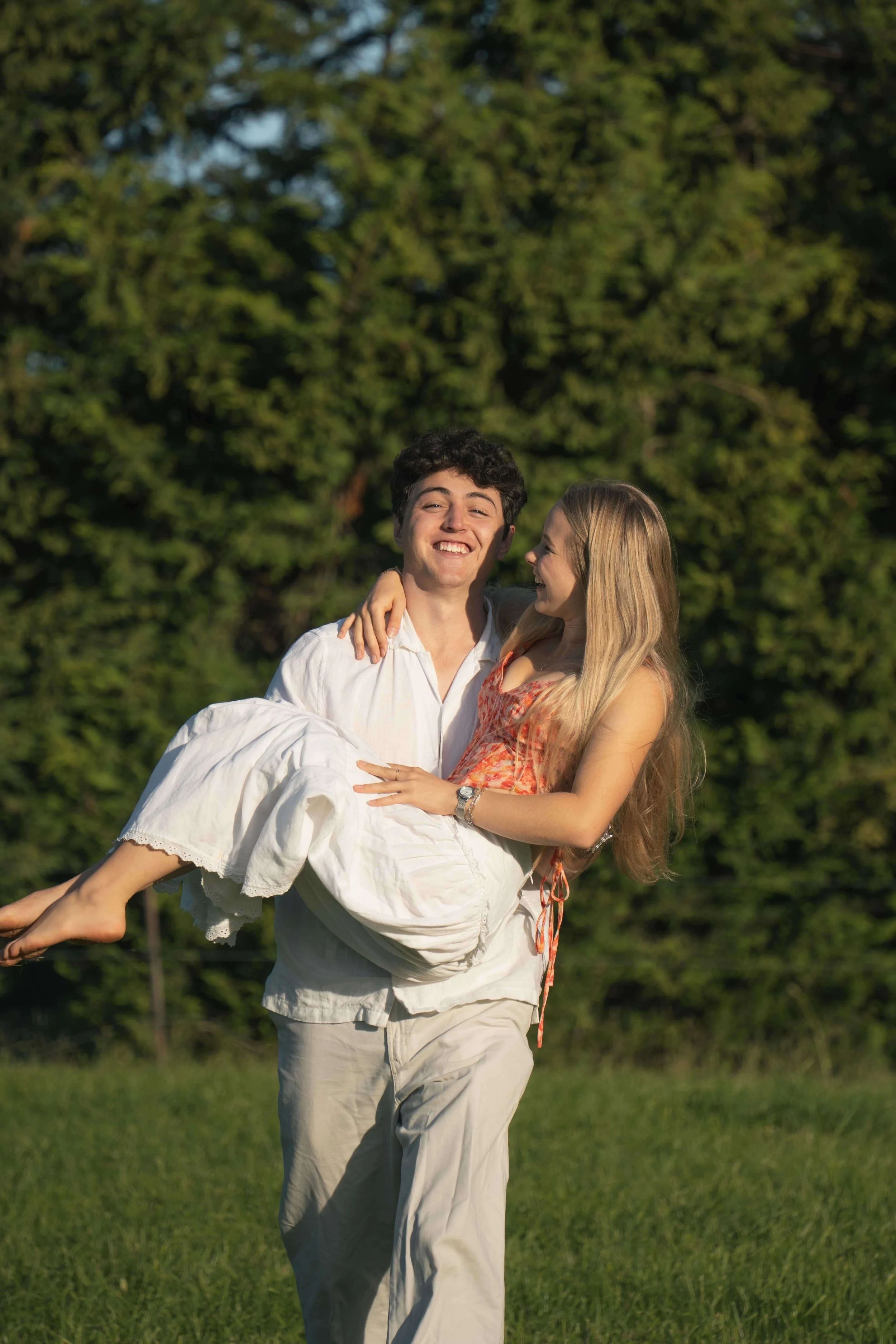A young man carrying a young woman in his arms outdoors with green trees in the background, both smiling and looking at each other.