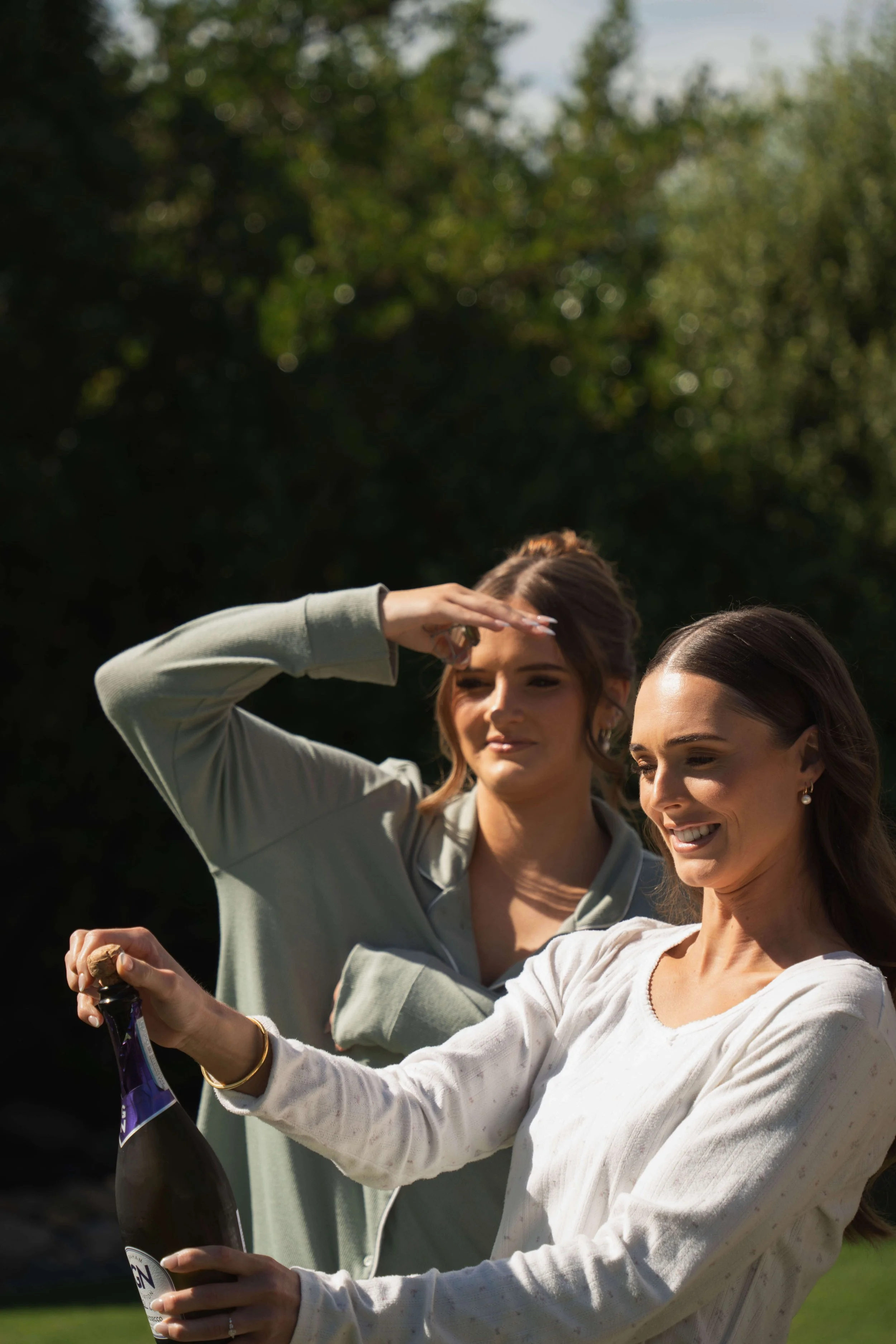 Two women outdoors, one opening a bottle of champagne while the other shields her eyes from the sun.